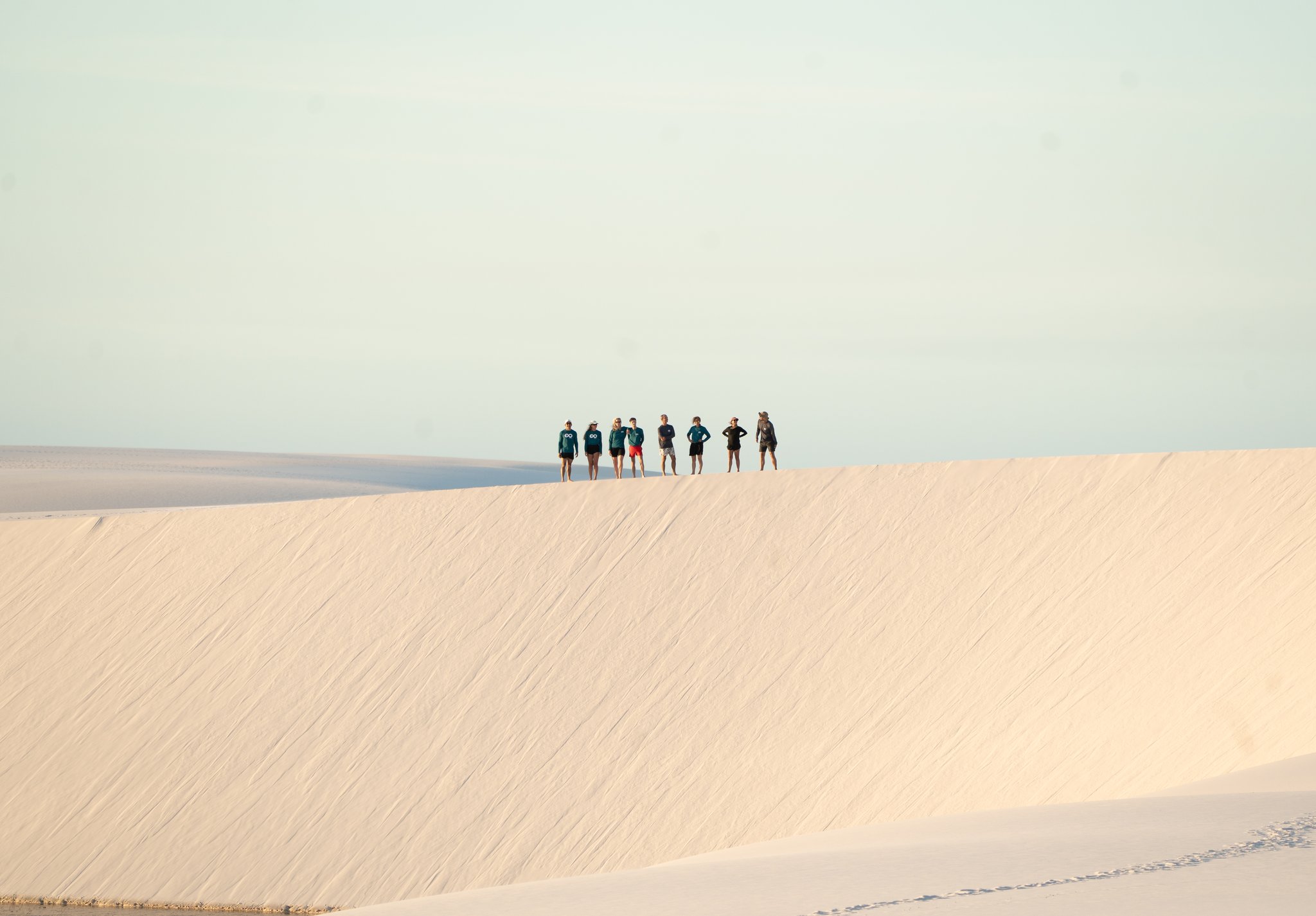 Seven figures, one immense ridge — the dunes put everything in perspective. A group of seven trekkers silhouetted along the crest of a vast white sand dune during a Lençóis Maranhenses trekking expedition, with pale sky and endless dunes stretching to the horizon