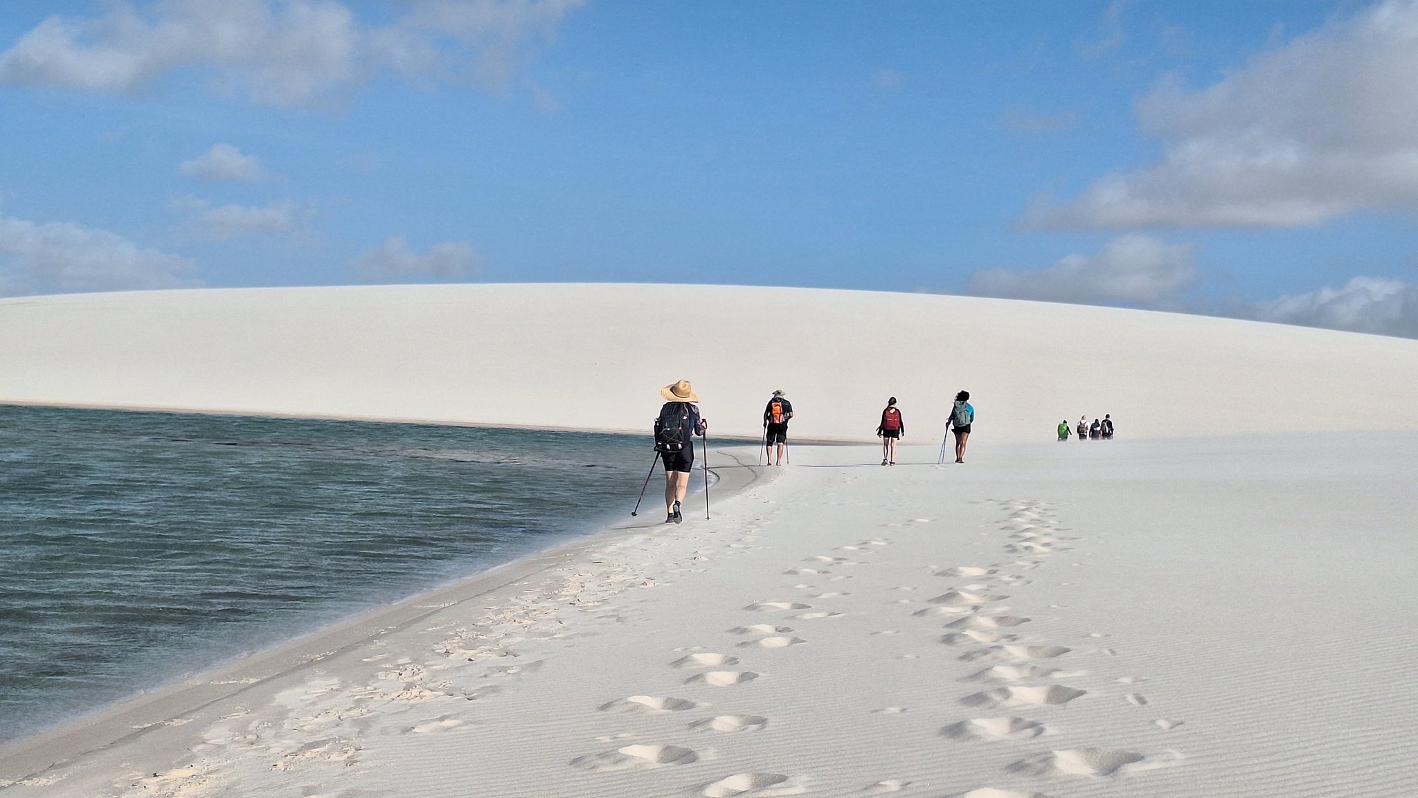 Single file along the ridge, the lagoon inches to one side and endless white dunes to the other. A group of hikers with trekking poles walks along a narrow white sand ridge beside a turquoise lagoon during a Lençóis Maranhenses trekking expedition, with a sweeping dune rising under a blue sky