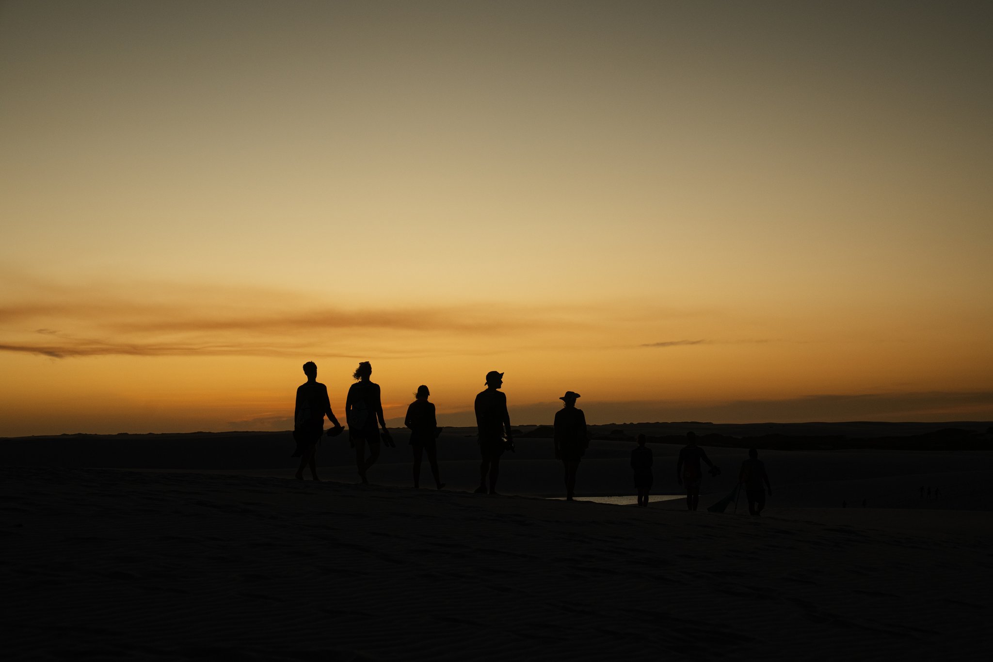 Eight silhouettes, one molten sky — the dunes at dusk belong entirely to them. Silhouettes of a group of trekkers walking across open dunes during a golden sunset on a Lençóis Maranhenses trekking expedition
