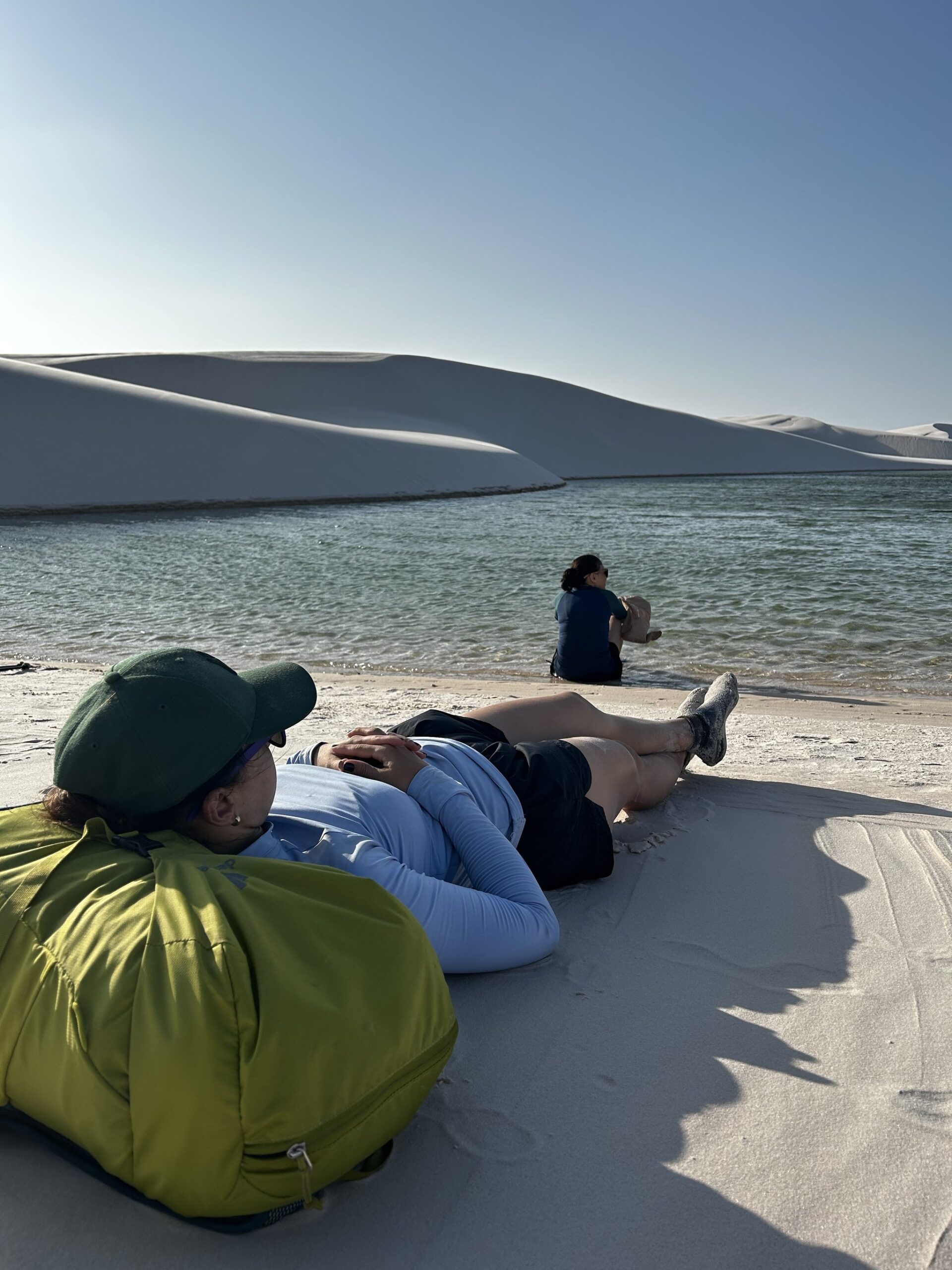 Cool water, warm sand — the perfect mid-trek pause. A trekker lies back on white sand dunes during a Lençóis Maranhenses trekking rest stop, while a companion sits at the edge of a clear turquoise lagoon with sweeping white dunes behind