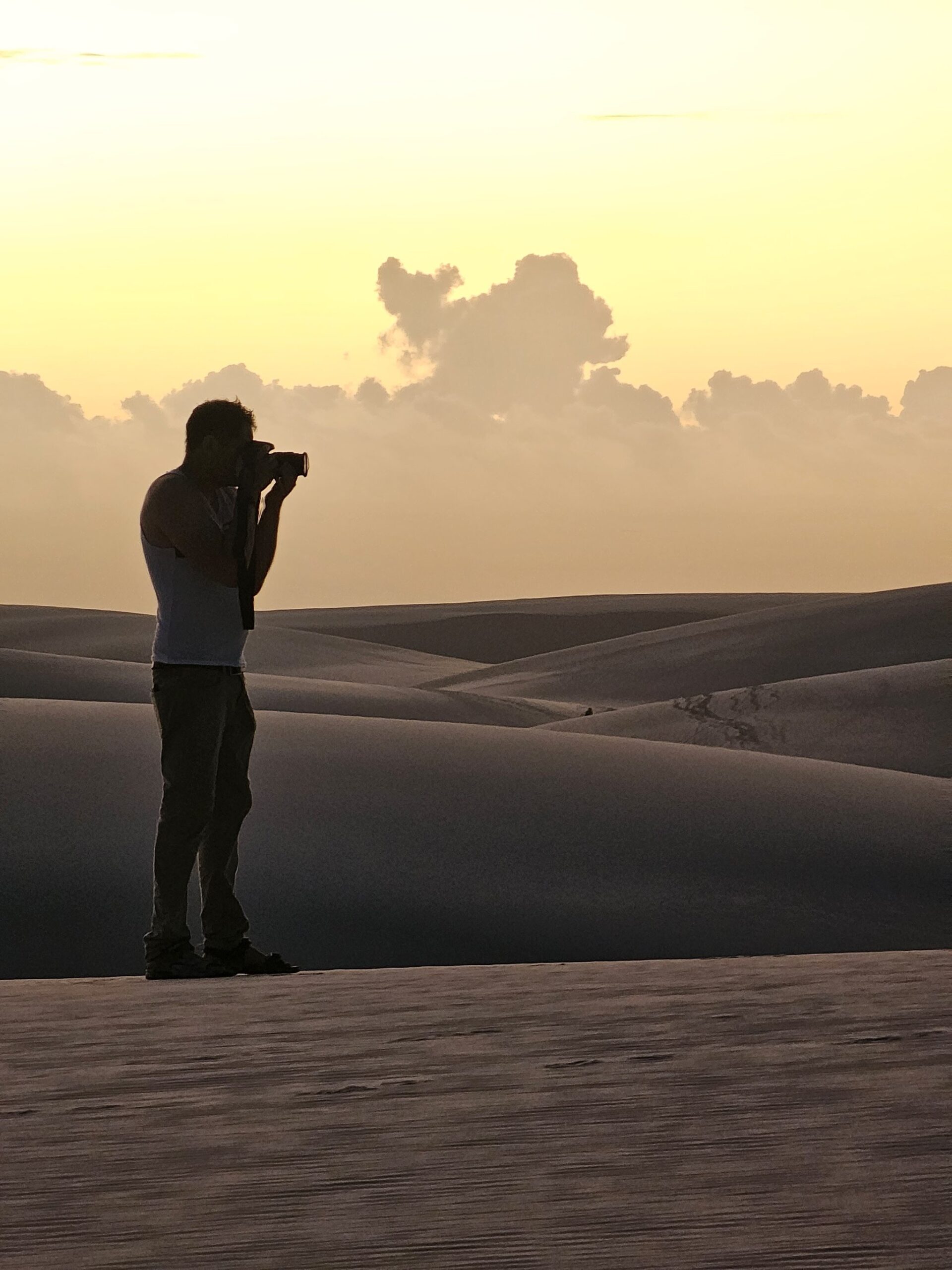 Silhouette of a photographer with a camera raised to their eye, standing on a white sand dune during a golden sunset over the rolling dune landscape of Lençóis Maranhenses trekking