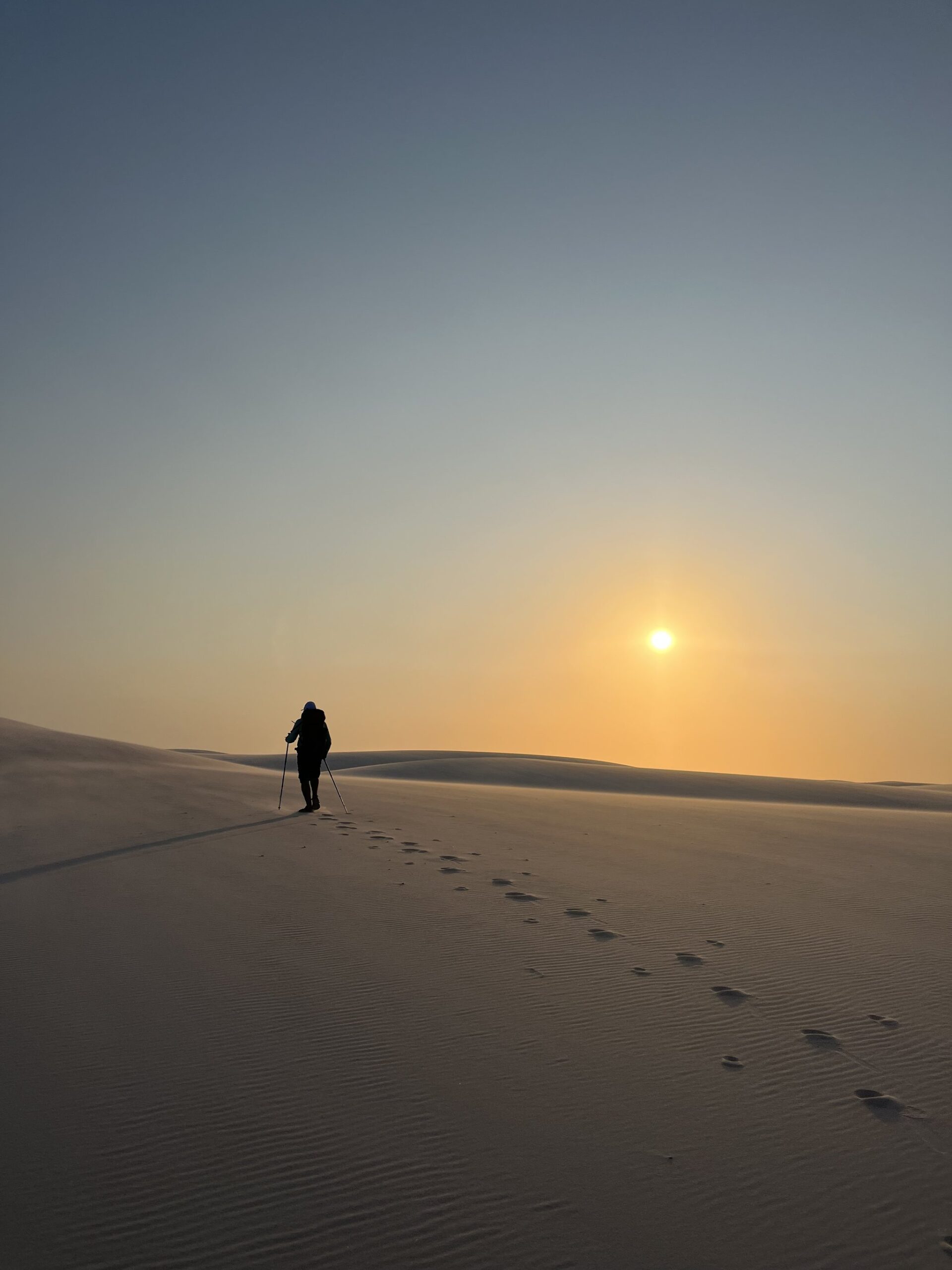 Just a silhouette, trekking poles, and the whole desert ahead. A lone trekker with hiking poles climbs a vast white sand dune toward a golden sunrise on a Lençóis Maranhenses trekking route, footprints trailing behind across the rippled sand