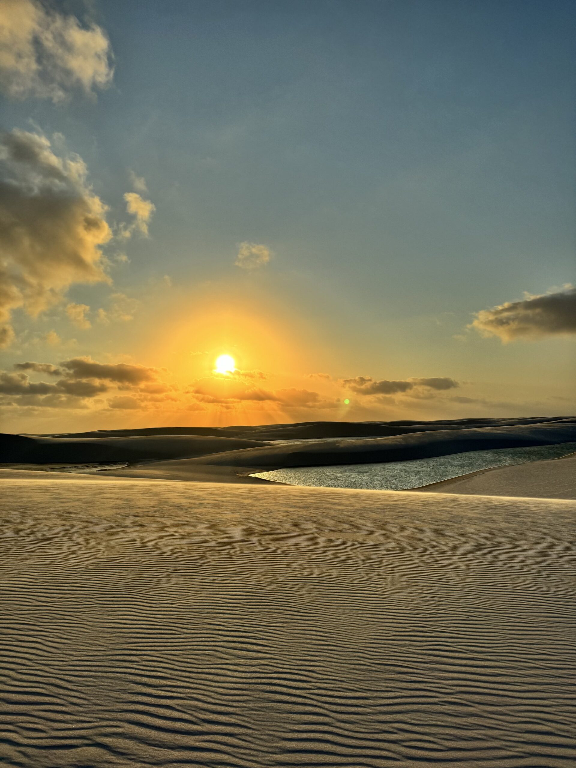 Dusk settles over the dunes, painting every ripple amber. Golden sunset over rippled white sand dunes with a still lagoon reflecting the sky during a Lençóis Maranhenses trekking crossing