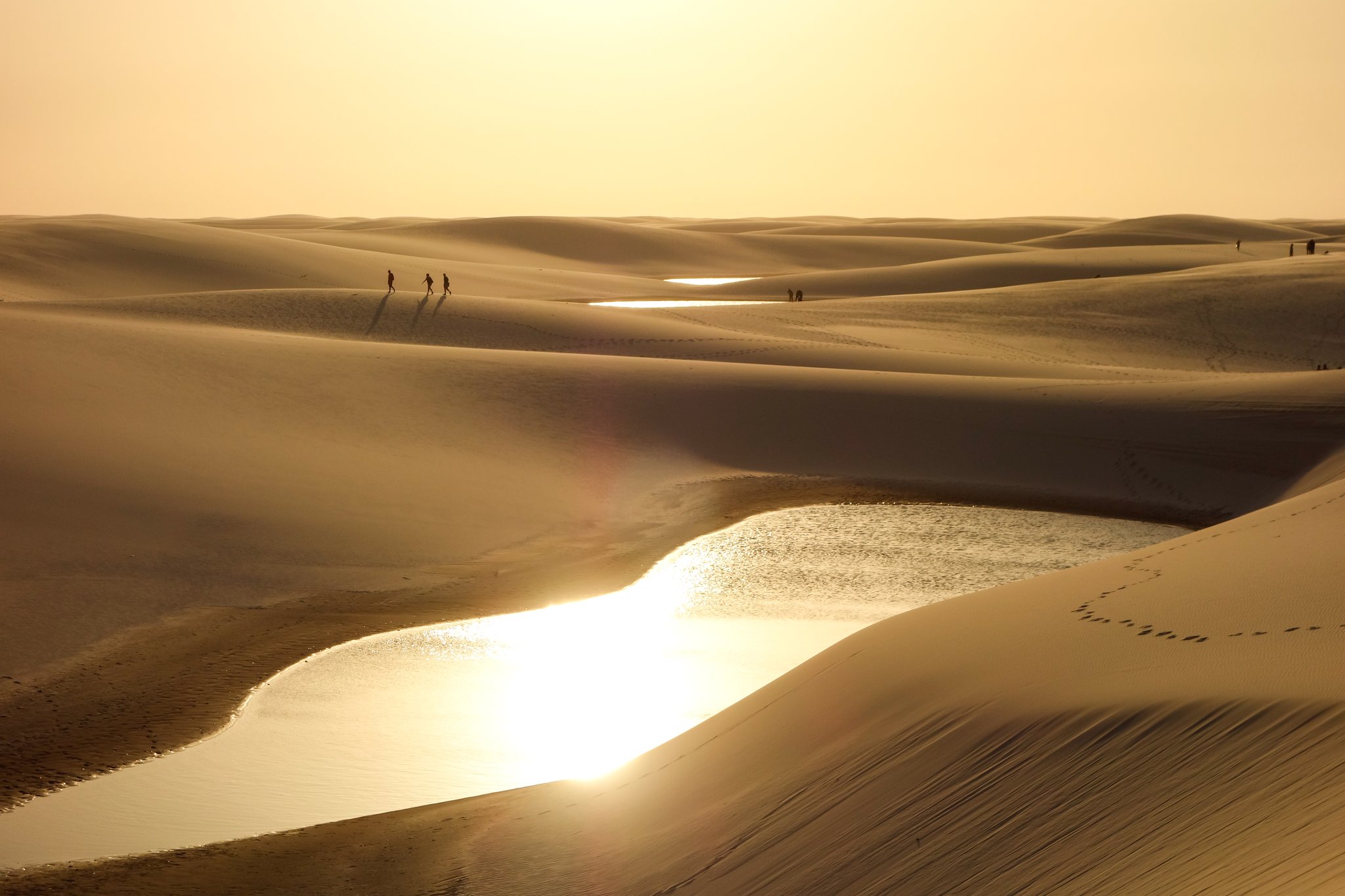 Silhouettes of trekkers walking across vast white sand dunes at golden hour, with shallow rainwater lagoons reflecting the sun during a Lençóis Maranhenses trekking route
