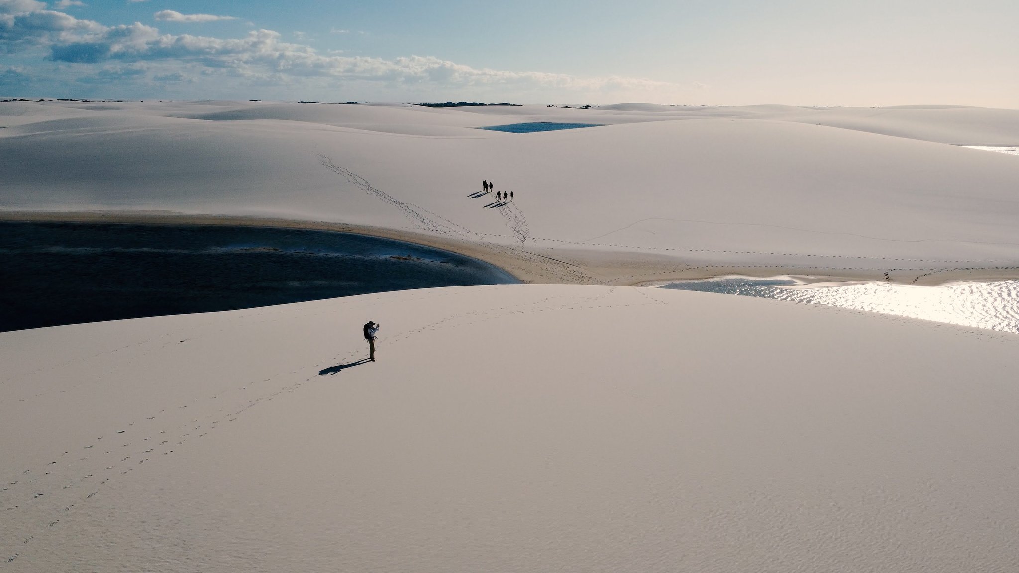 Aerial view of a lone hiker and a small group walking across vast white dunes beside a dark lagoon on a Lençóis Maranhenses trip, their footprints tracing separate paths through the sand