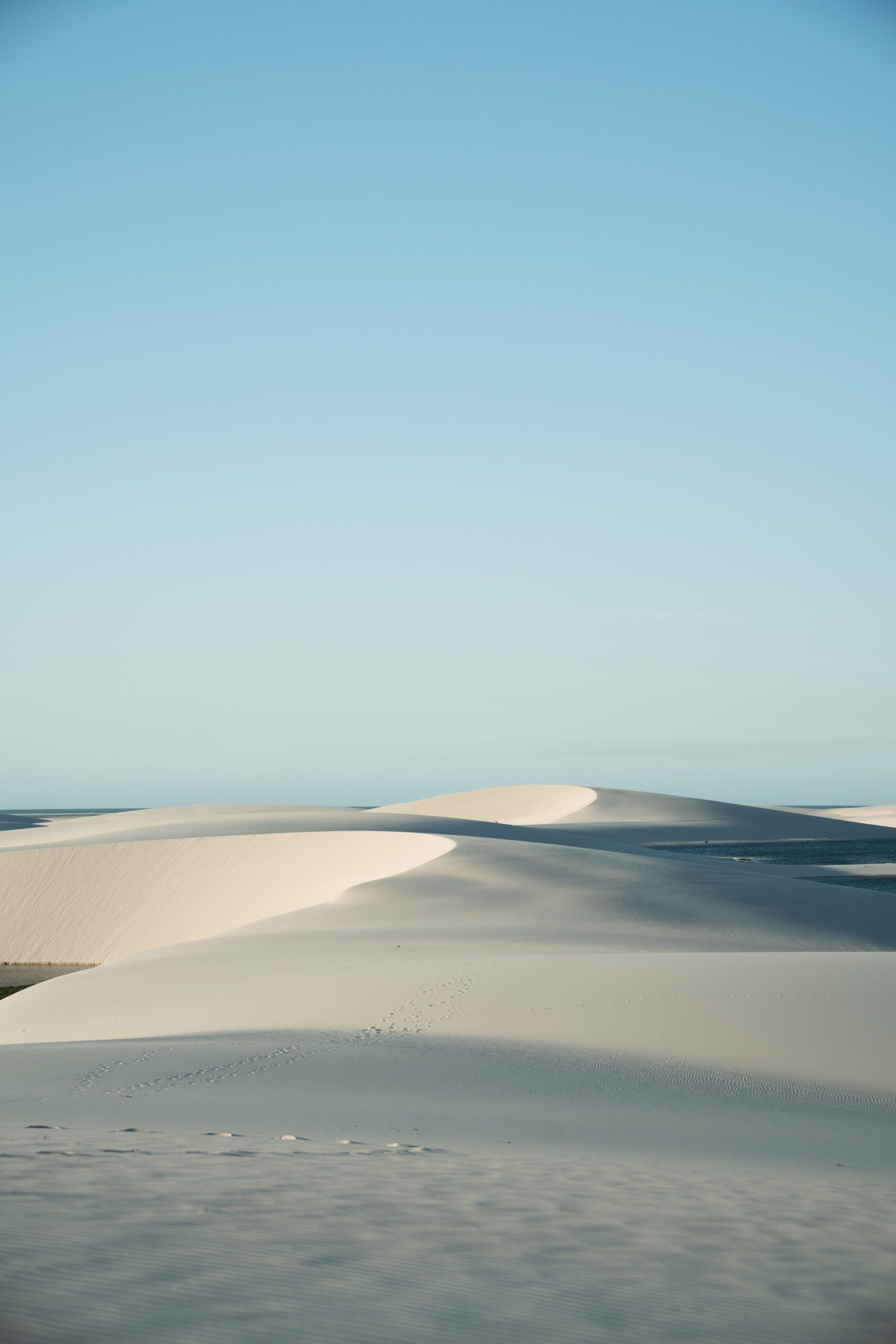 Sweeping white sand dunes under a pale blue sky on a Lençóis Maranhenses trip, with a faint trail of footprints climbing the nearest slope