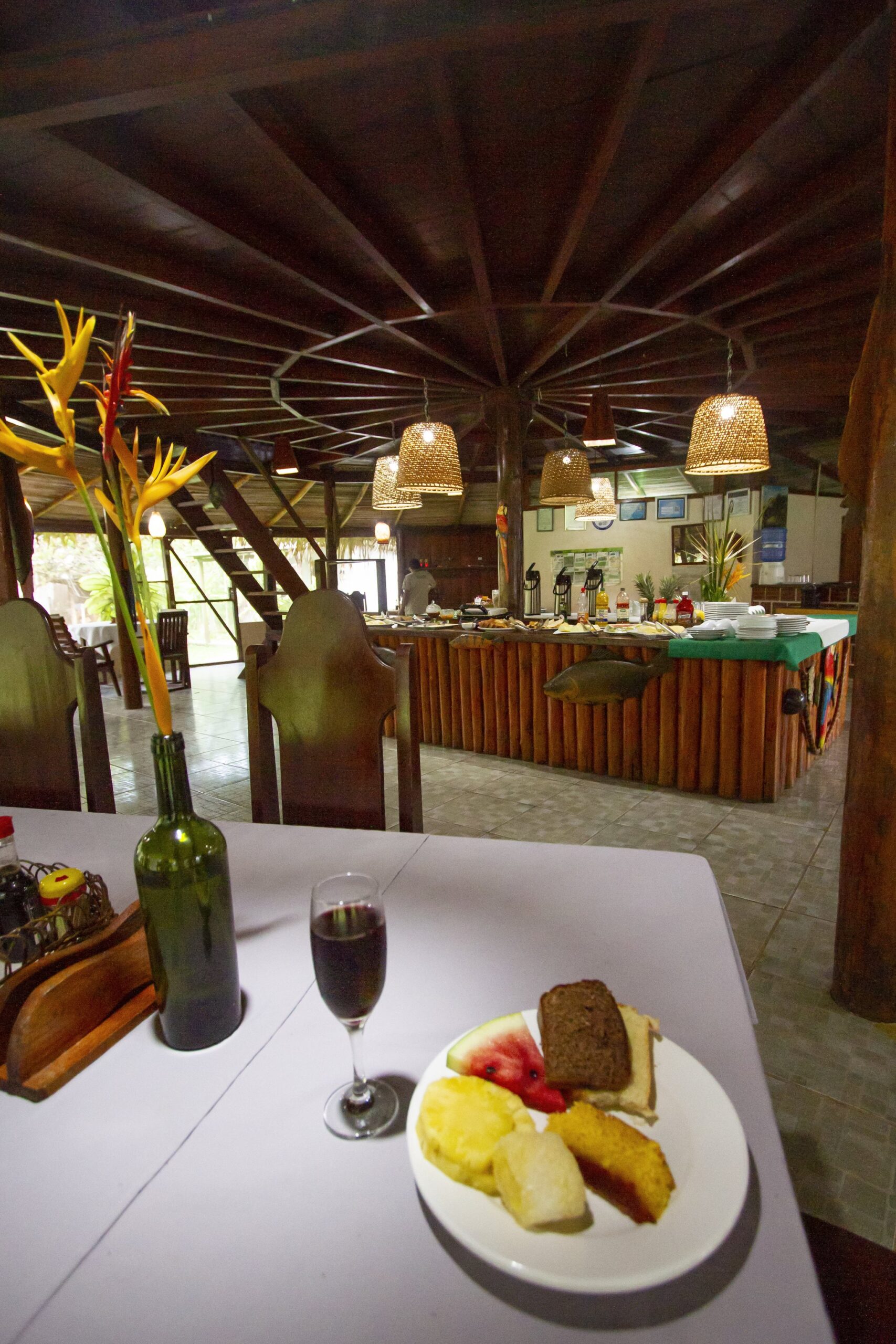 Welcome plate and glass of wine on a table inside a wooden Amazon lodge dining hall with buffet beyond