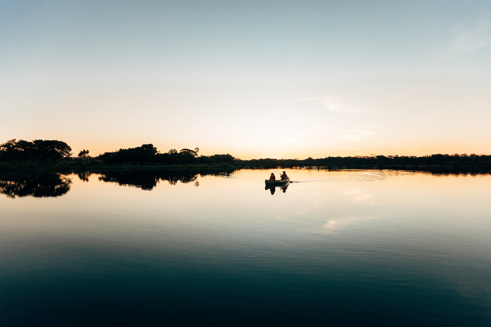 At dusk the river turns to glass, a single canoe drifting across the silence. Photo: Felipe Castellari Lone canoe with two paddlers silhouetted on a mirror-calm river at dusk on an Amazon rainforest vacation in Brazil