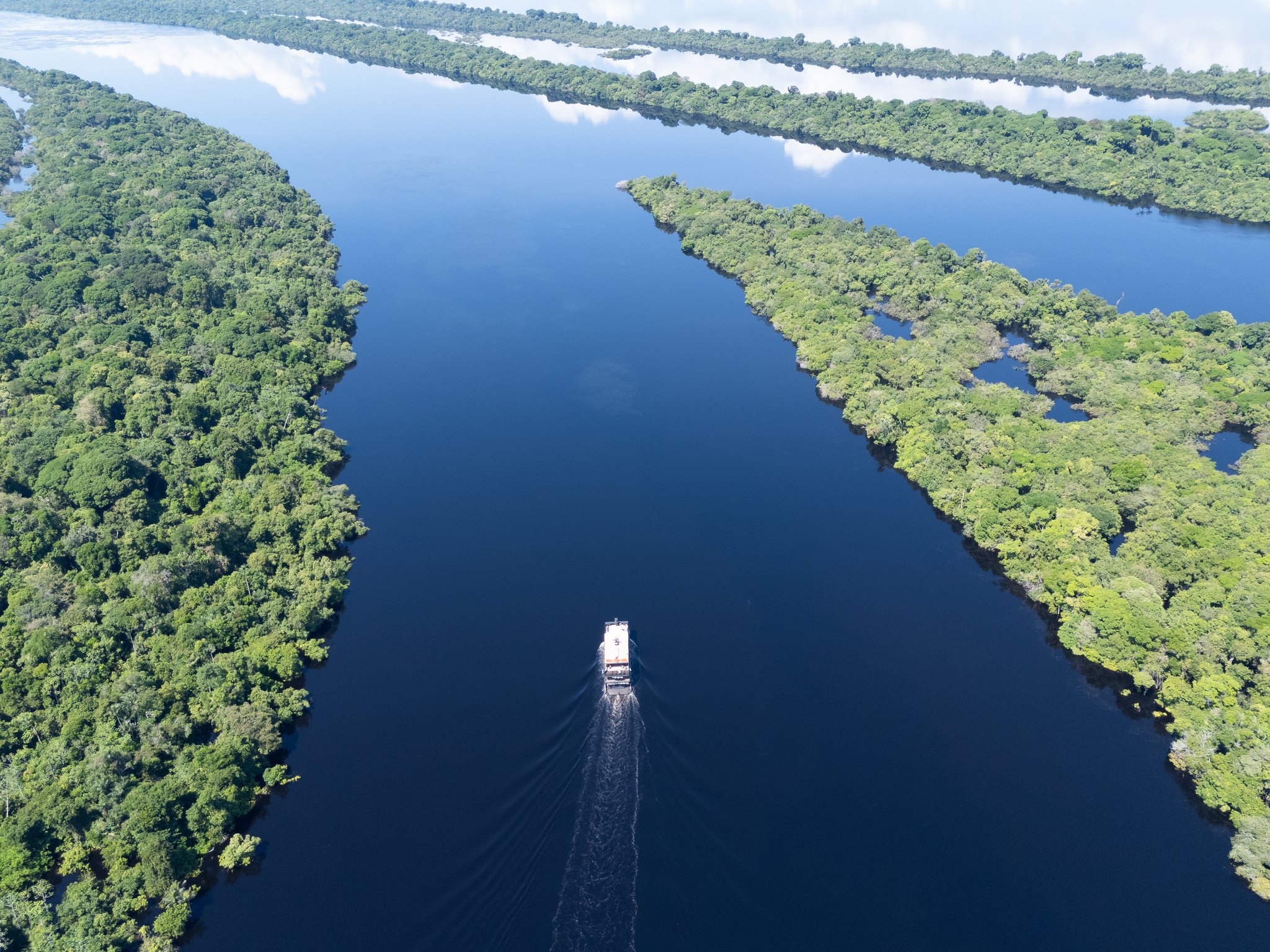 Aerial view of a riverboat navigating a dark-water channel flanked by dense Amazon rainforest canopy on a luxury Amazon cruise in Brazil