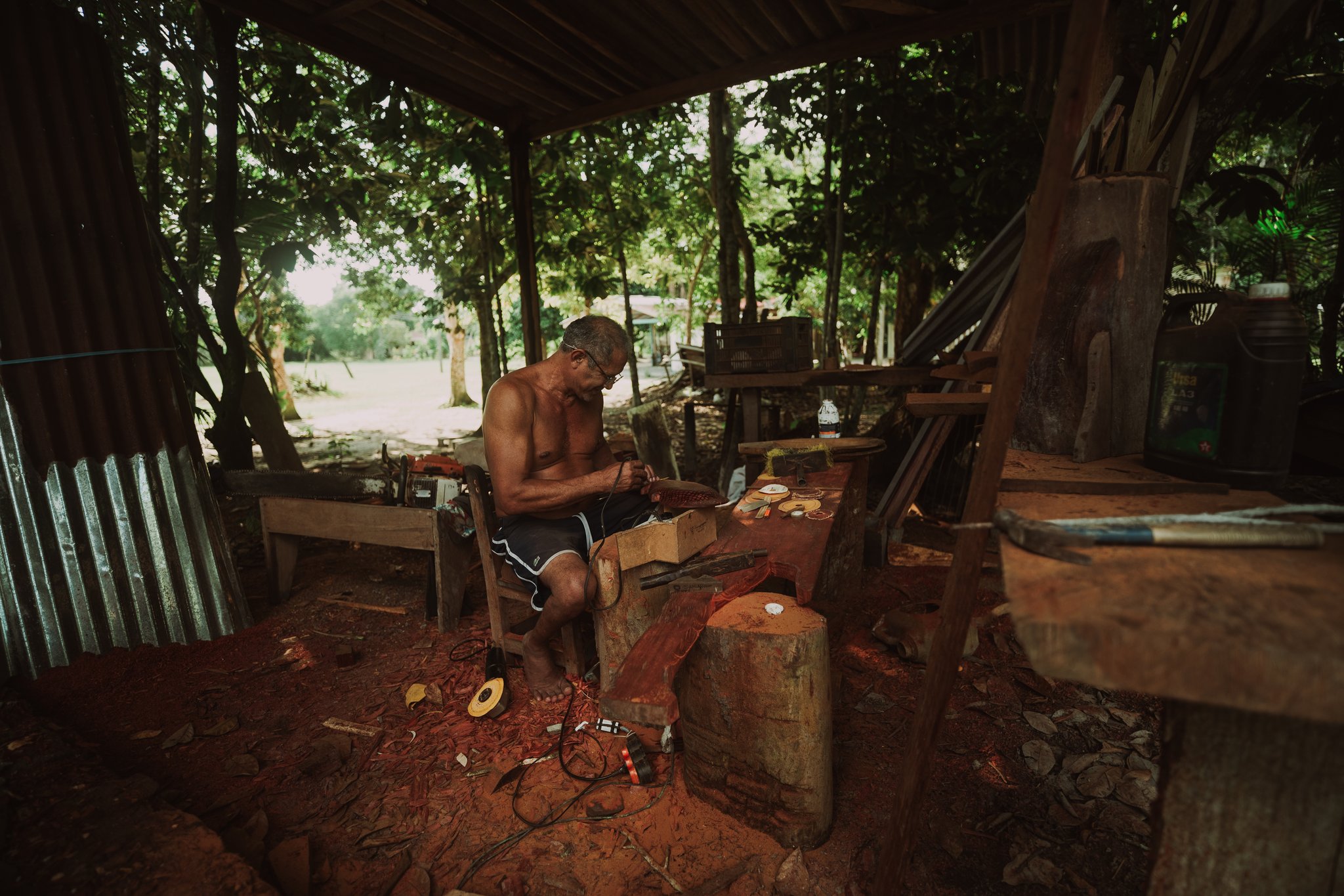 A local craftsman sits shirtless in an open-air riverside workshop, carefully hand-finishing a wooden piece surrounded by tools and red-earth soil on a Luxury Amazon Cruise in Brazil community visit
