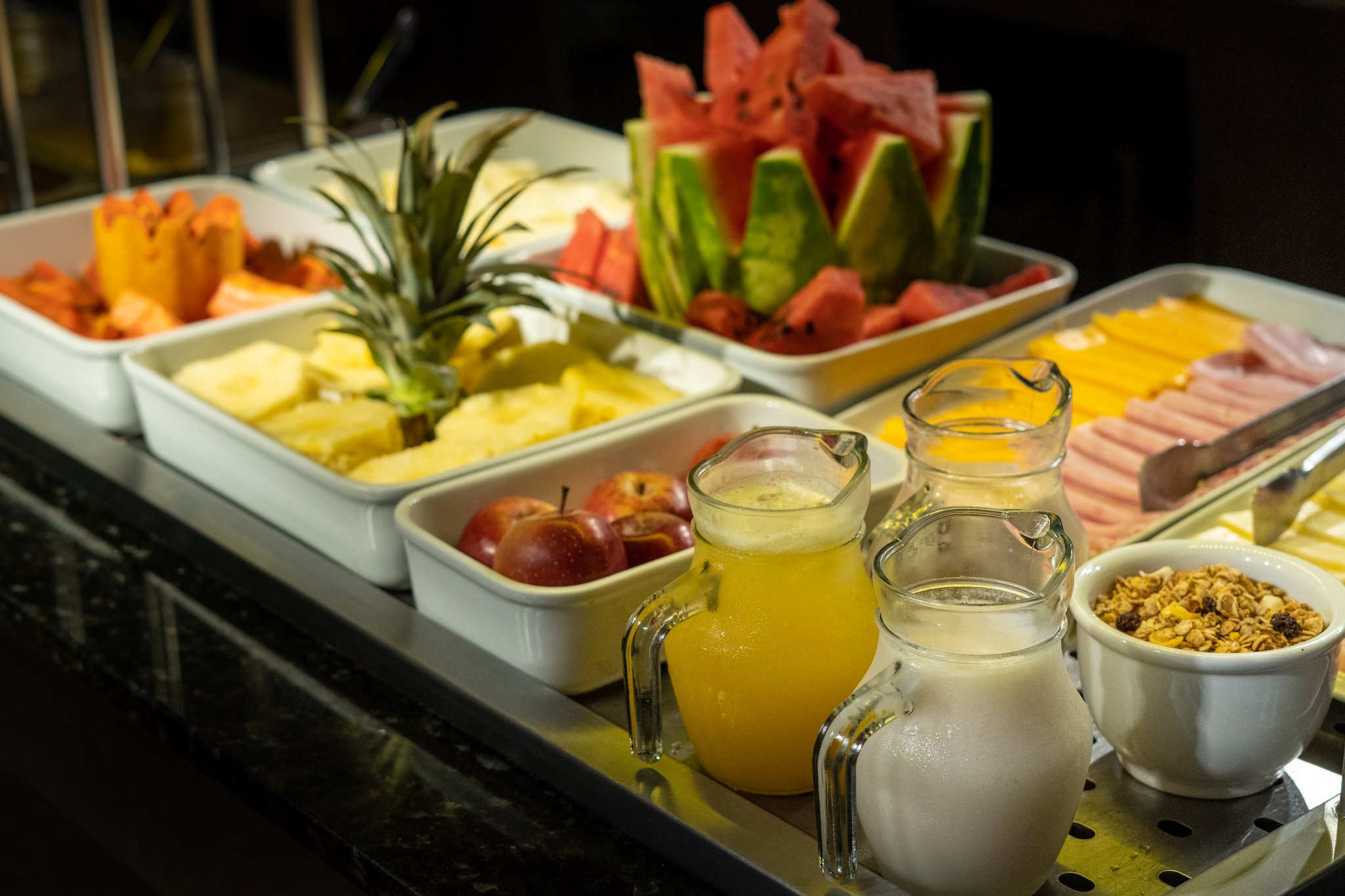 Um farto buffet de café da manhã a bordo de um cruzeiro de luxo pelo Amazonas no Brasil, com bandejas de abacaxi, melancia, maçãs e mamão fatiados, jarras de suco natural, leite, frios, queijo e uma tigela de granola