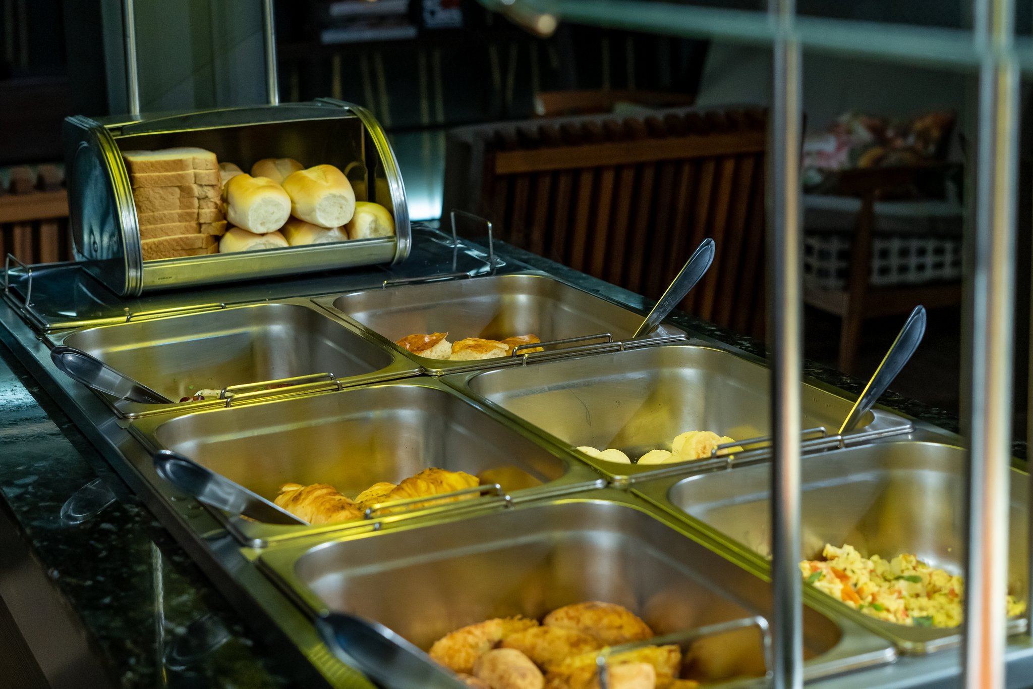 A warm breakfast buffet spread aboard a luxury Amazon cruise in Brazil, featuring golden pastries, bread rolls, sliced sandwich bread, scrambled eggs, and a vegetable rice dish in polished stainless-steel chafing trays