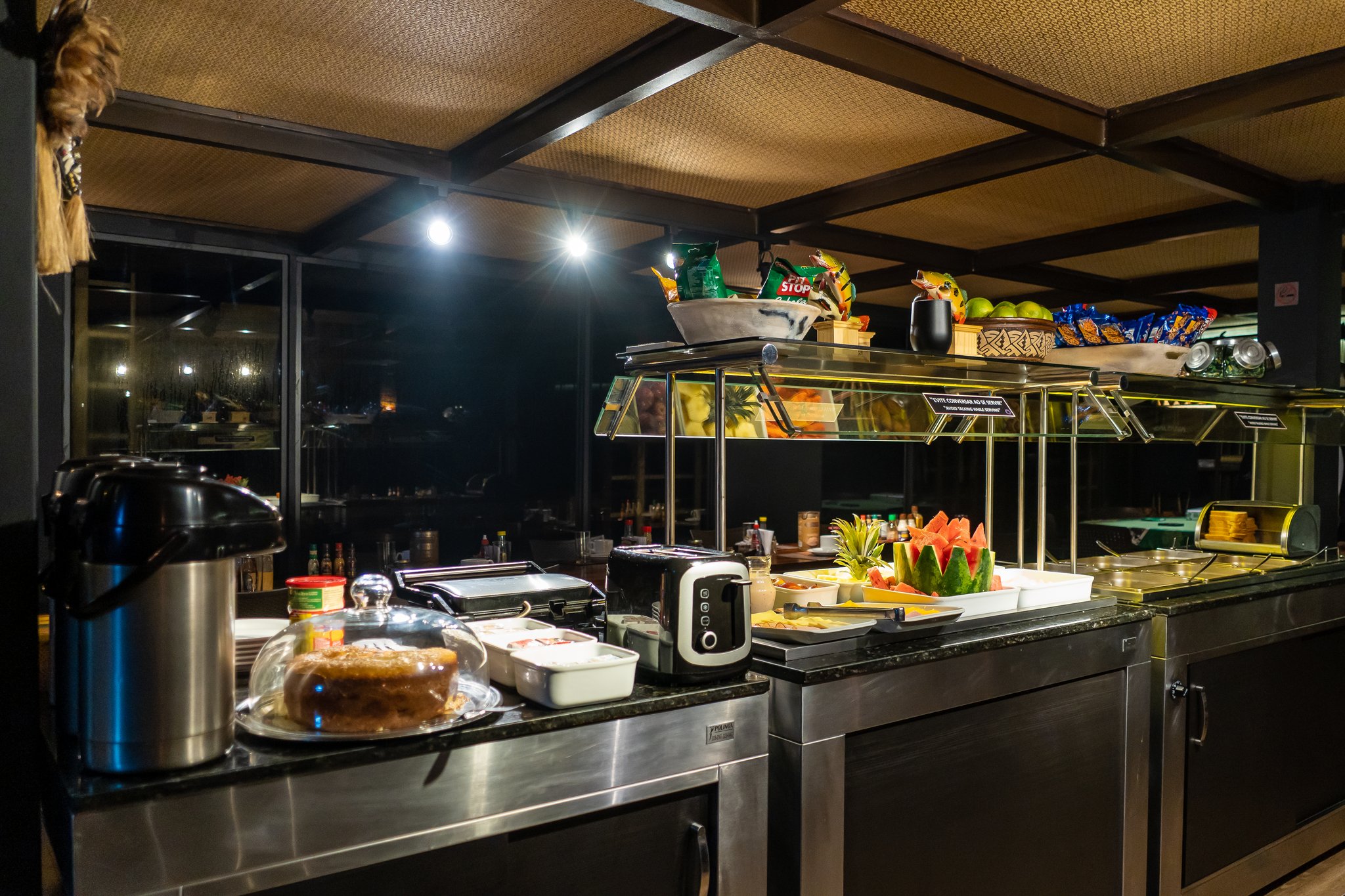 Buffet iluminado a bordo de um cruzeiro de luxo na Amazônia brasileira, com melancia fatiada, frutas tropicais frescas, um bolo inteiro sob redoma de vidro, uma torradeira, garrafa térmica de café e lanches em prateleiras escalonadas, com vista noturna para o rio através de grandes janelas