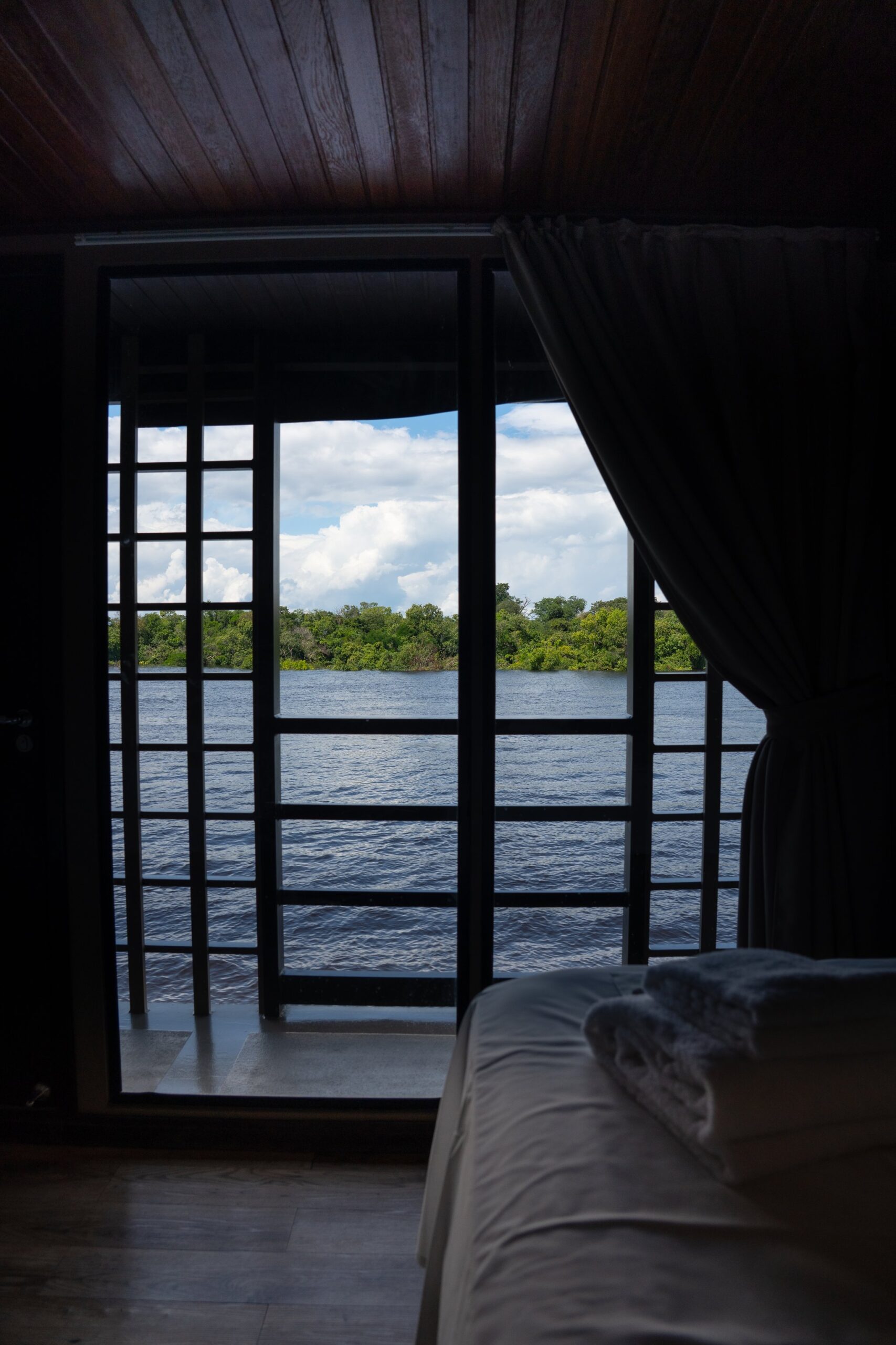 Vista de dentro de uma cabine com revestimento de madeira escura a bordo de um cruzeiro de luxo na Amazônia brasileira — janelas gradeadas do piso ao teto emolduram um rio largo e mata verde sob céu nublado