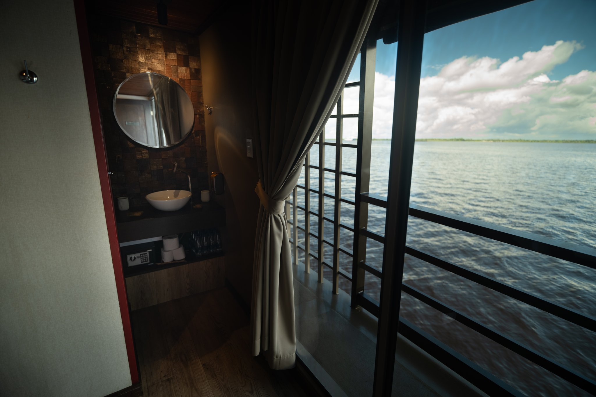 Interior de suíte a bordo de um cruzeiro de luxo pela Amazônia no Brasil, com espelho redondo, cuba de apoio, cofre e janelas do piso ao teto com vista para o rio amplo