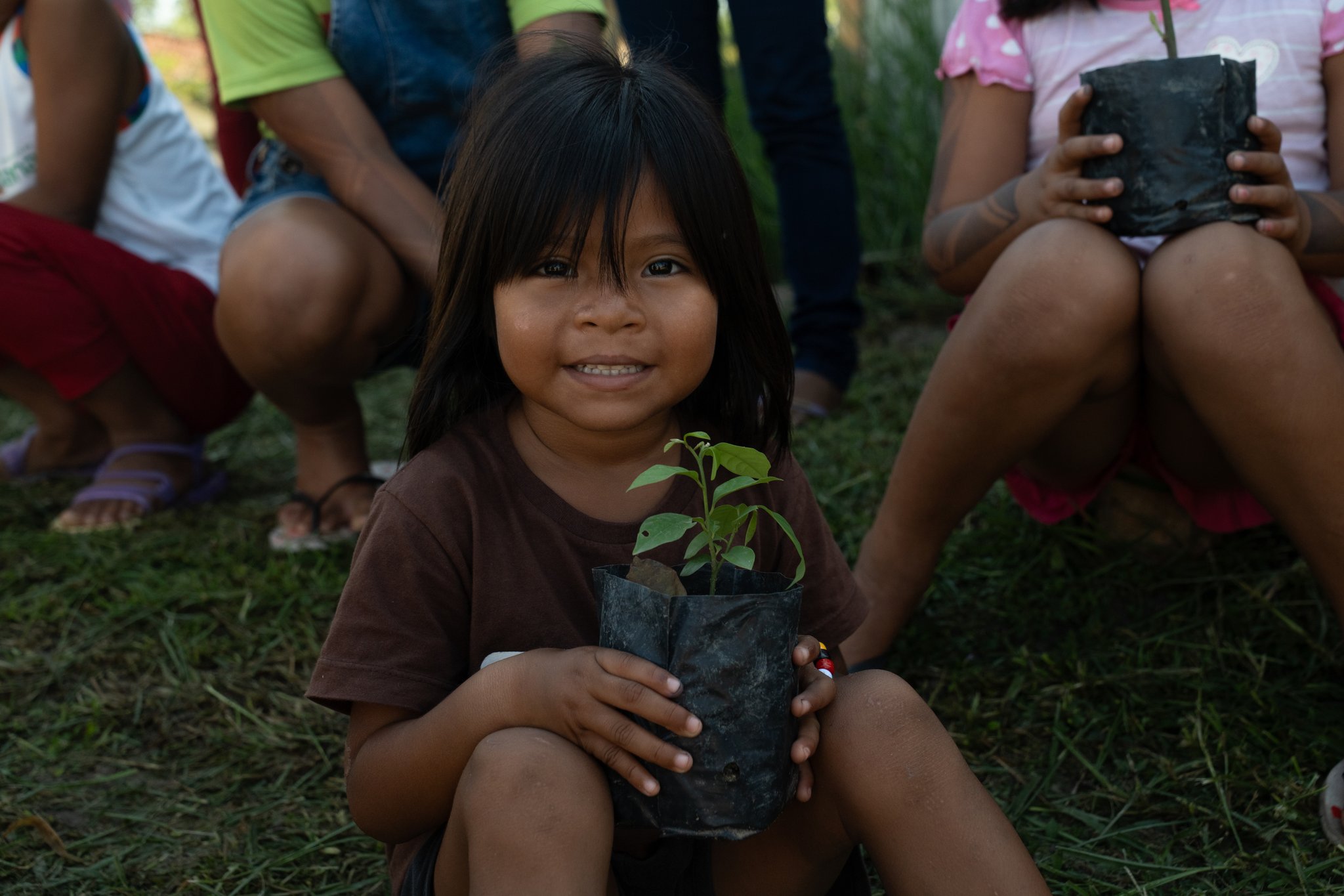 Uma menina sorridente segura uma muda verde em um saquinho preto durante uma visita comunitária em um cruzeiro de luxo pela Amazônia no Brasil, com outras crianças sentadas na grama ao fundo