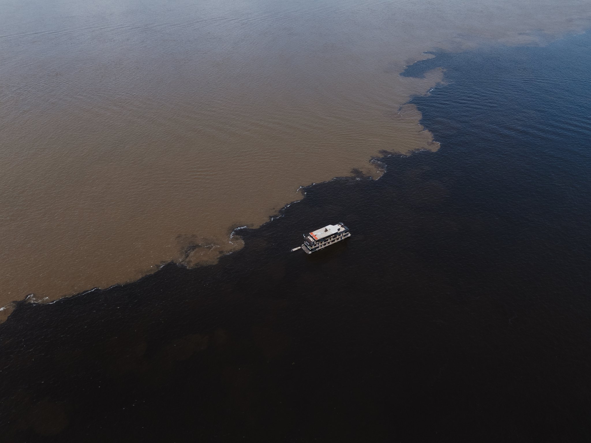 Vista aérea de um barco fluvial cruzando o Encontro das Águas na Amazônia, onde as águas negras e as águas barrentas se encontram em uma fronteira sinuosa e contrastante