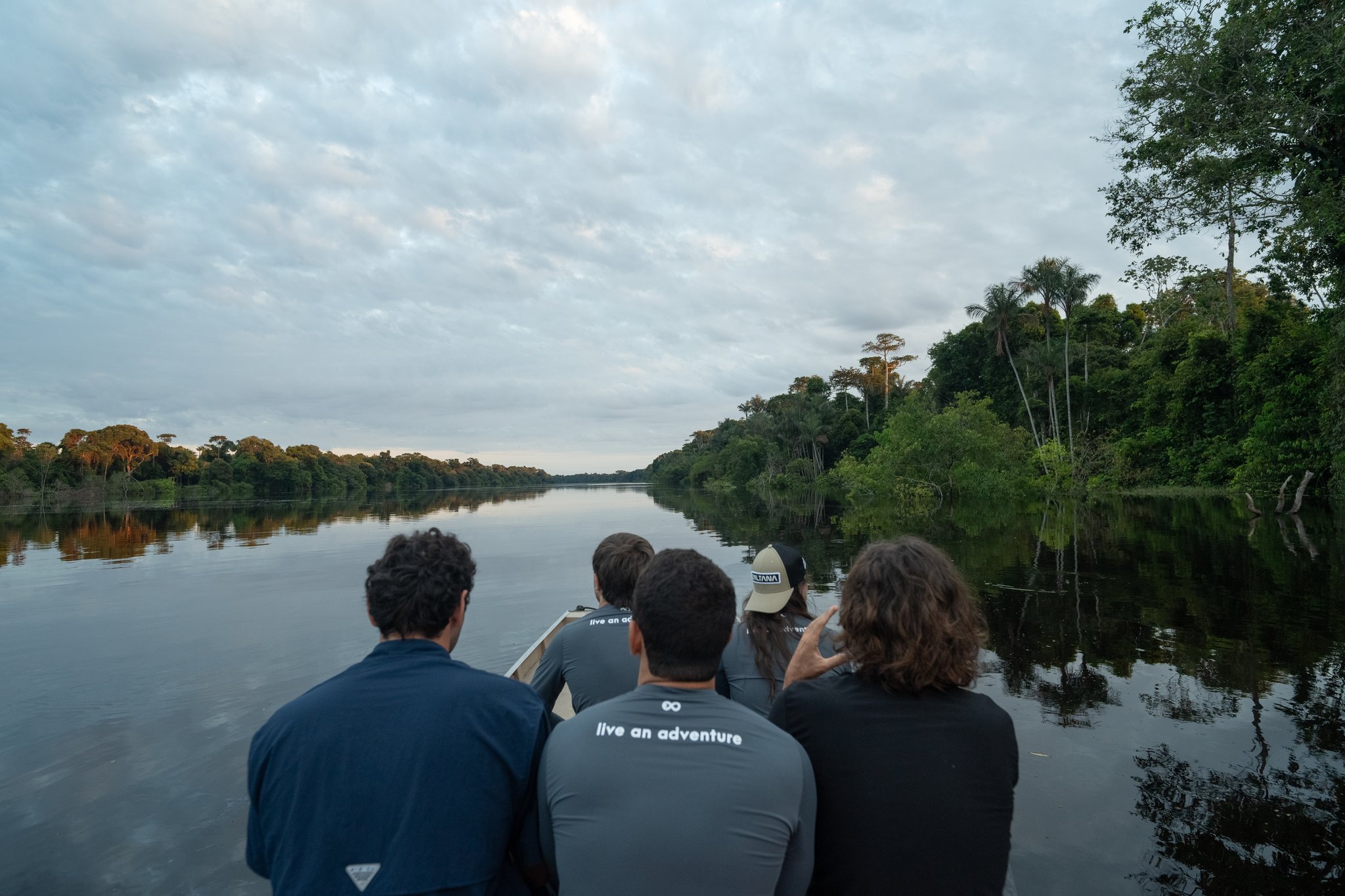 Quatro viajantes sentados na proa de um barco pequeno num rio de águas escuras e calmas, com floresta densa em ambas as margens, durante um cruzeiro de luxo pelo Amazonas no Brasil