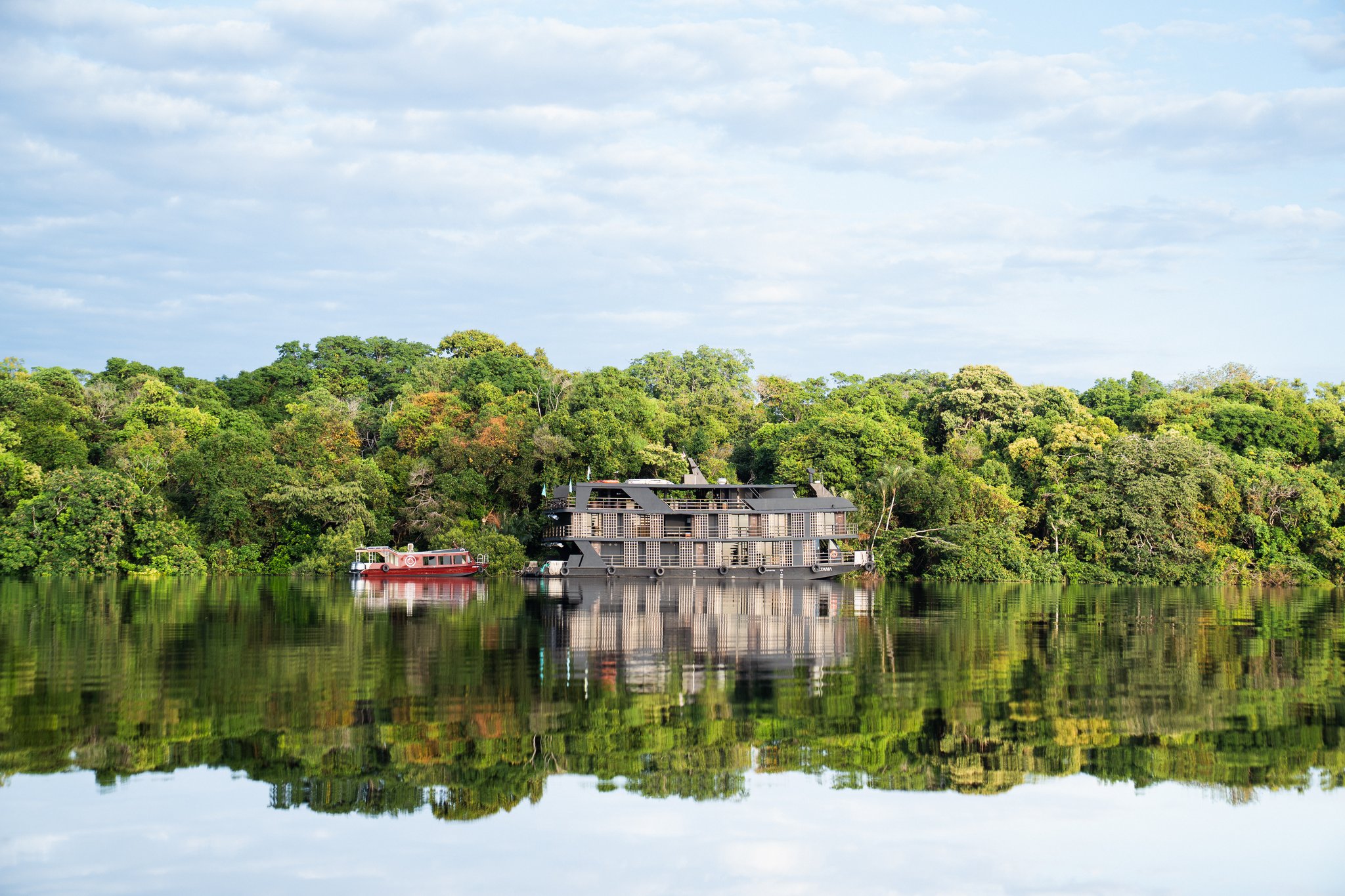 A multi-deck luxury Amazon cruise riverboat moored along a lush tropical forest riverbank, with perfect mirror reflections on the calm dark water and a red support boat alongside