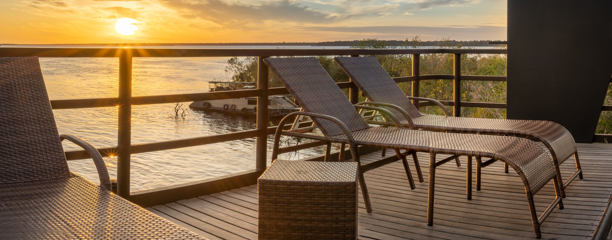 Espreguiçadeiras de vime em deck de madeira com vista para um rio largo ao entardecer dourado durante um cruzeiro de luxo na Amazônia brasileira