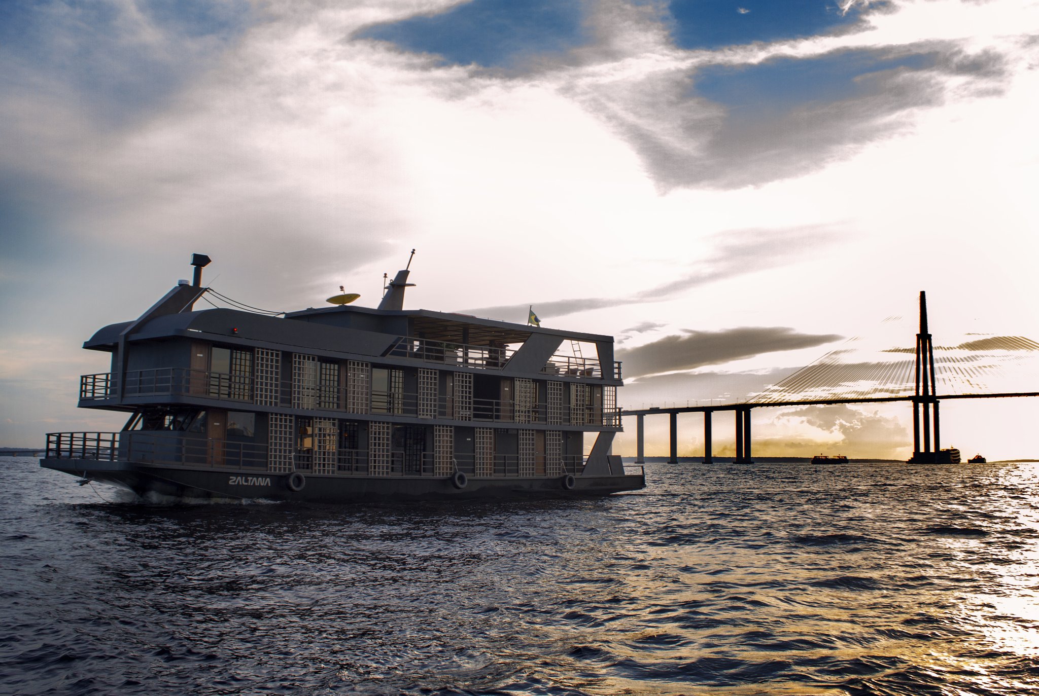 O barco fluvial Zaltana partindo para um cruzeiro de luxo na Amazônia brasileira, com uma ponte estaiada em silhueta contra um pôr do sol dramático sobre o largo Rio Negro