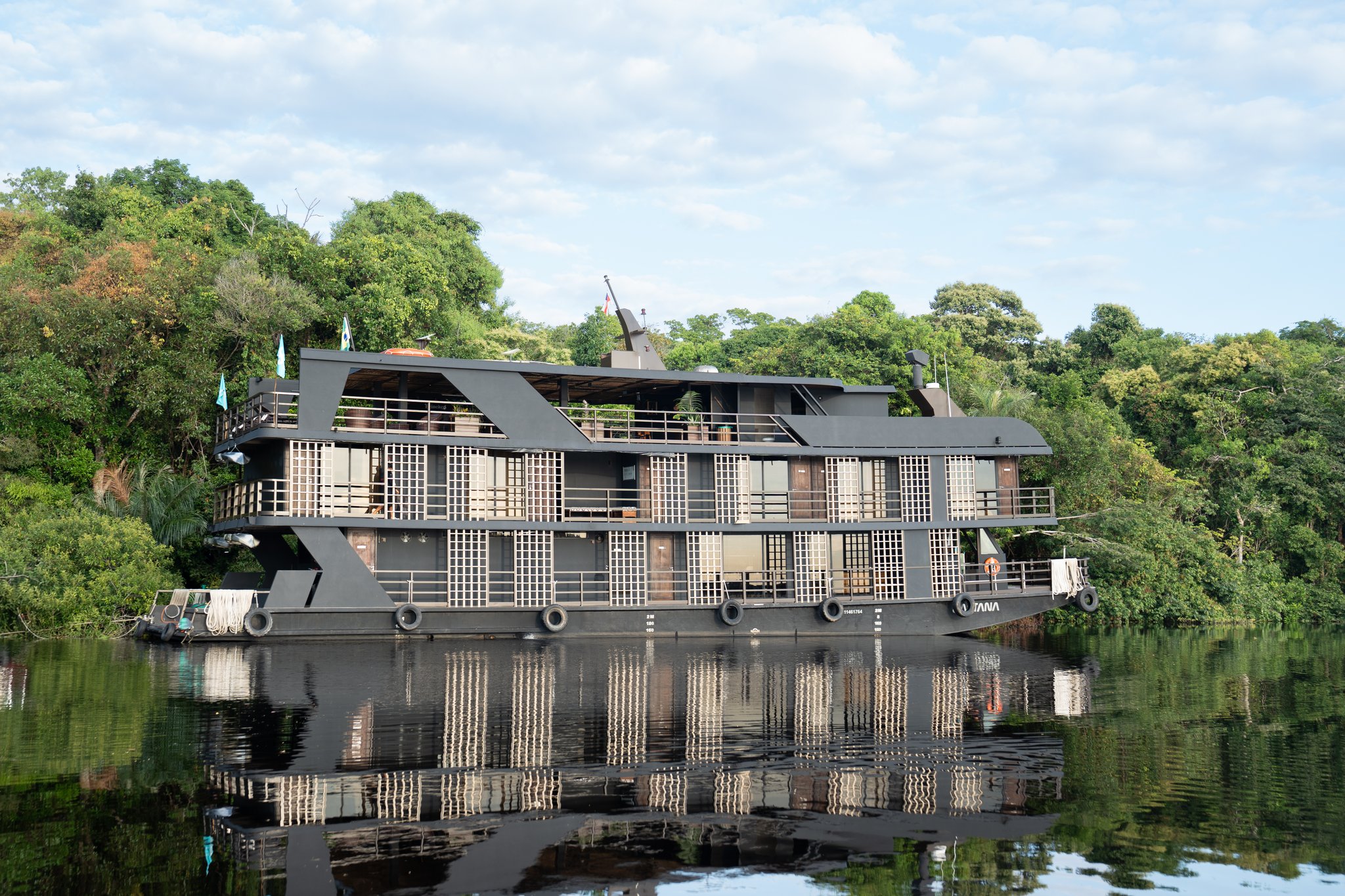O barco de cruzeiro de luxo Zaltana ancorado em um rio de águas negras completamente calmas, com seu casco escuro de múltiplos decks e cabines com grades refletidas na superfície, ladeado por densa floresta amazônica