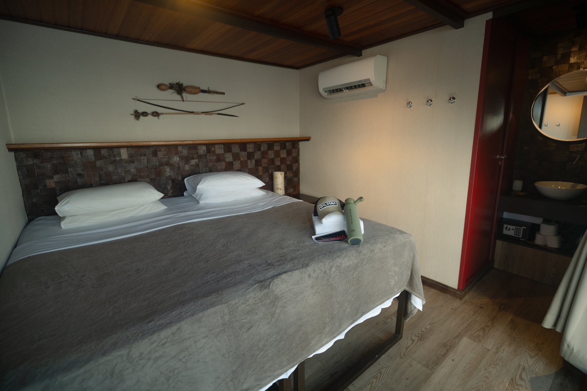 Cama de casal arrumada com travesseiros brancos e manta cinza em uma suíte do barco Zaltana, com cabeceira em mosaico de madeira, decoração de arco e flechas indígenas na parede, teto de madeira e ar-condicionado
