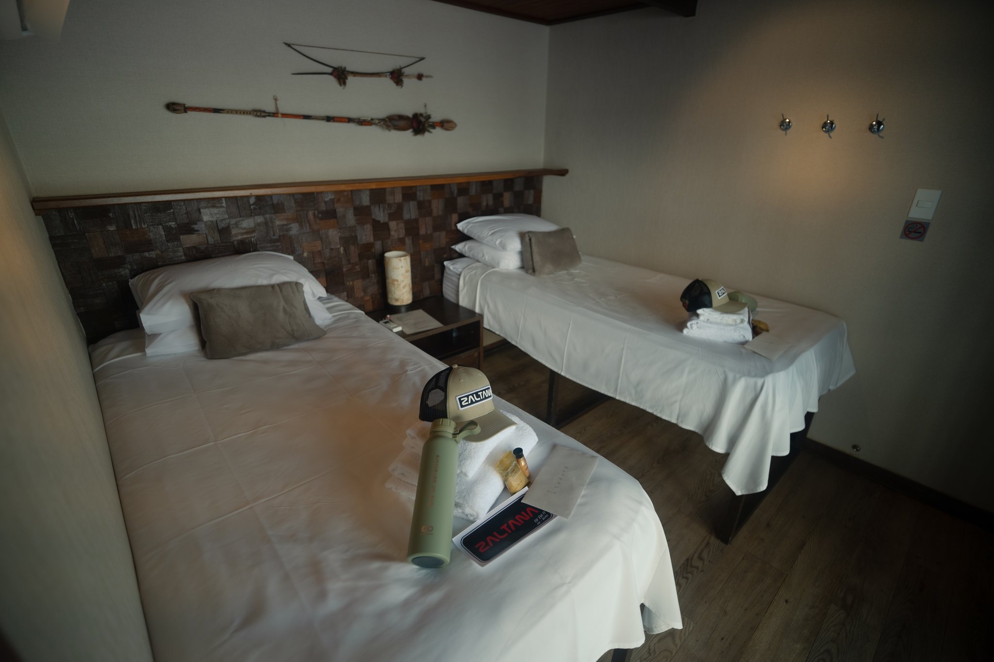 Cabine dupla a bordo do barco fluvial Zaltana em um Luxury Amazon Cruise in Brazil, com roupas de cama brancas impecáveis, cabeceira em mosaico de pedra, artefatos decorativos indígenas na parede e amenidades da Zaltana sobre as camas