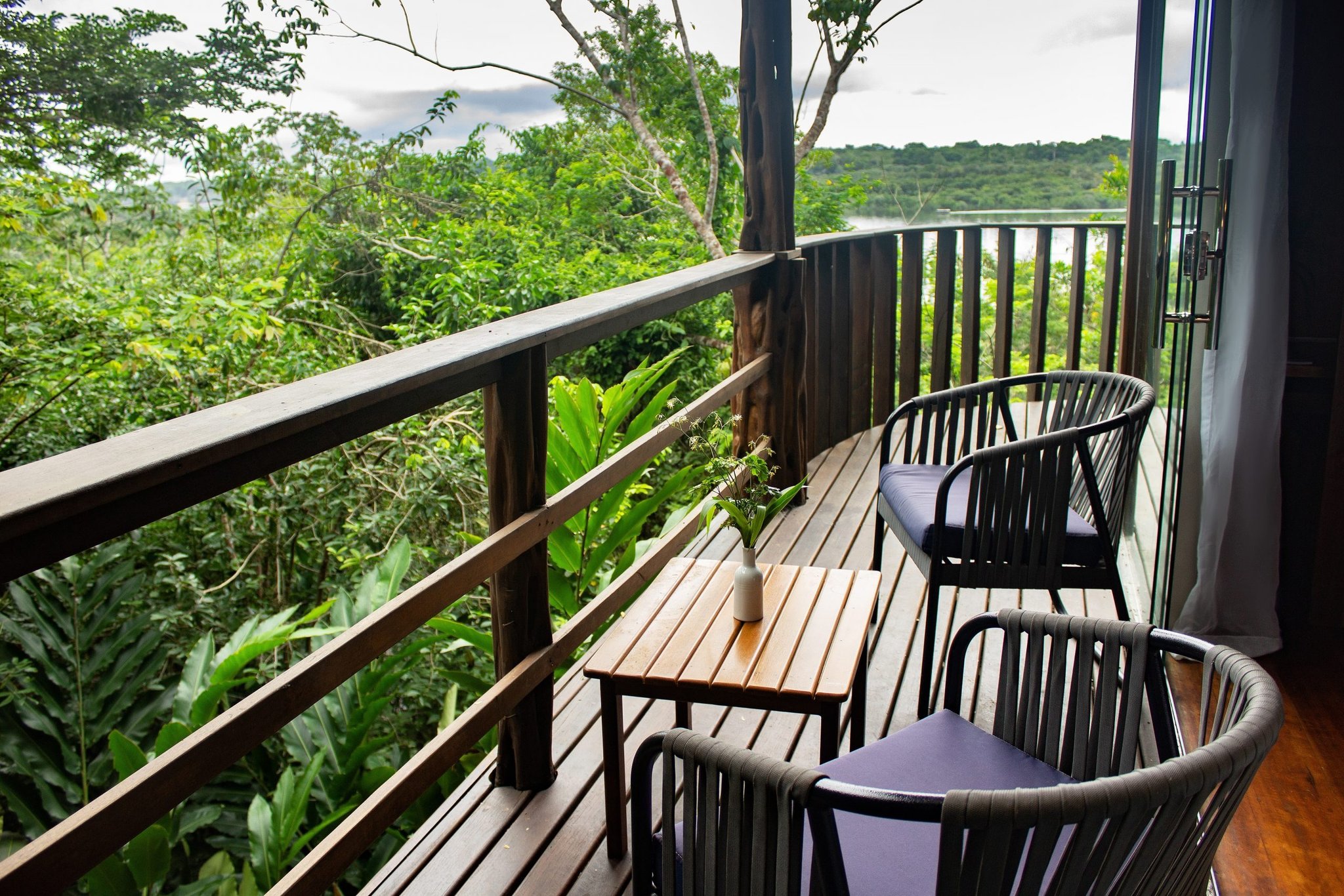 Two chairs, a forest stretching to the horizon — mornings don't get quieter than this. A curved wooden balcony at a luxury amazon lodge brazil with two rattan chairs and a small teak table overlooking dense green rainforest canopy and a distant river