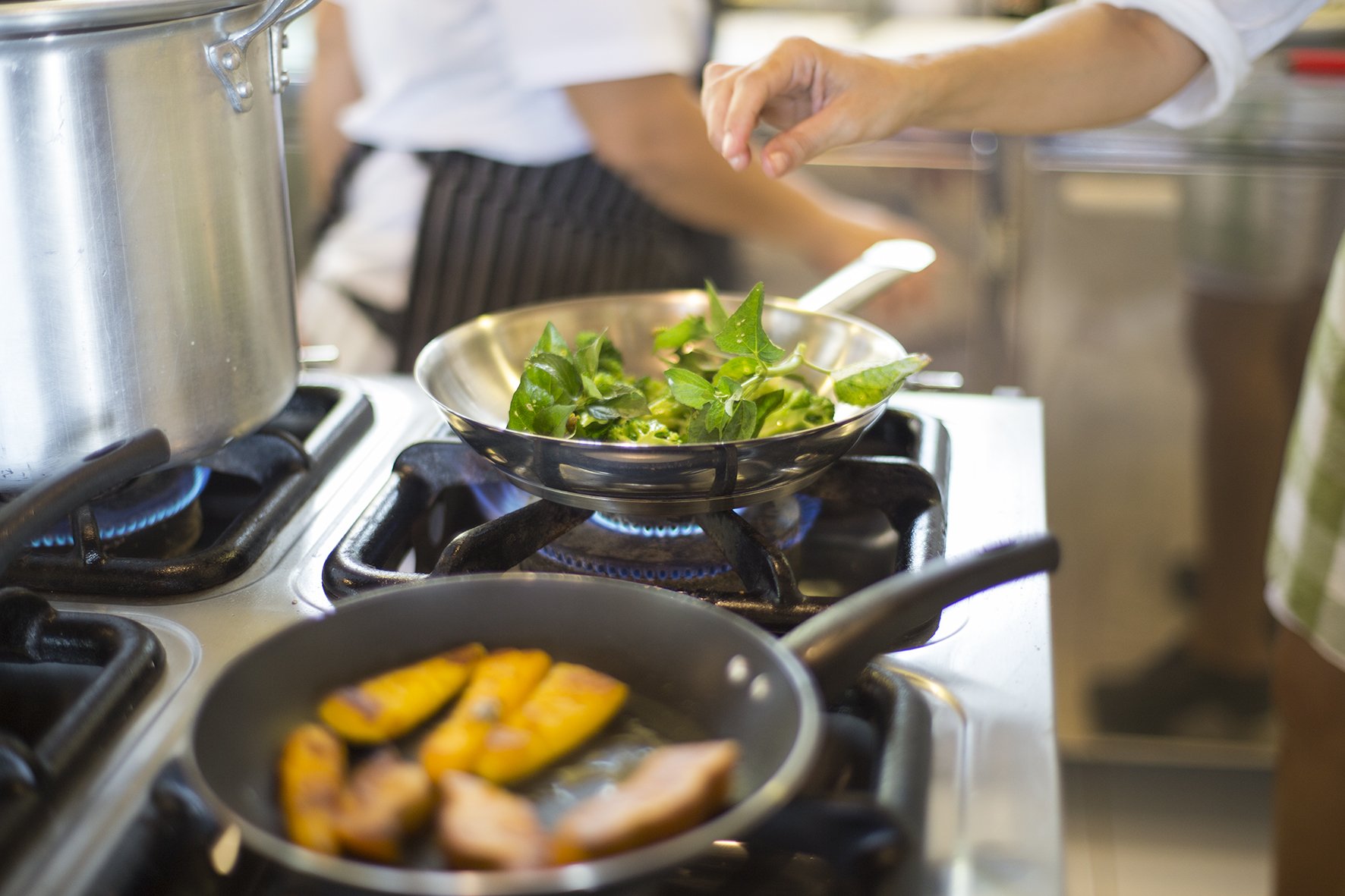 Duas frigideiras, uma chama — sabores da floresta ganhando vida. Um chef de avental listrado salteia folhas verdes frescas em uma frigideira de aço inox sobre chama a gás na cozinha de um lodge de luxo na Amazônia brasileira, com fatias de fruta douradas em uma segunda frigideira em primeiro plano
