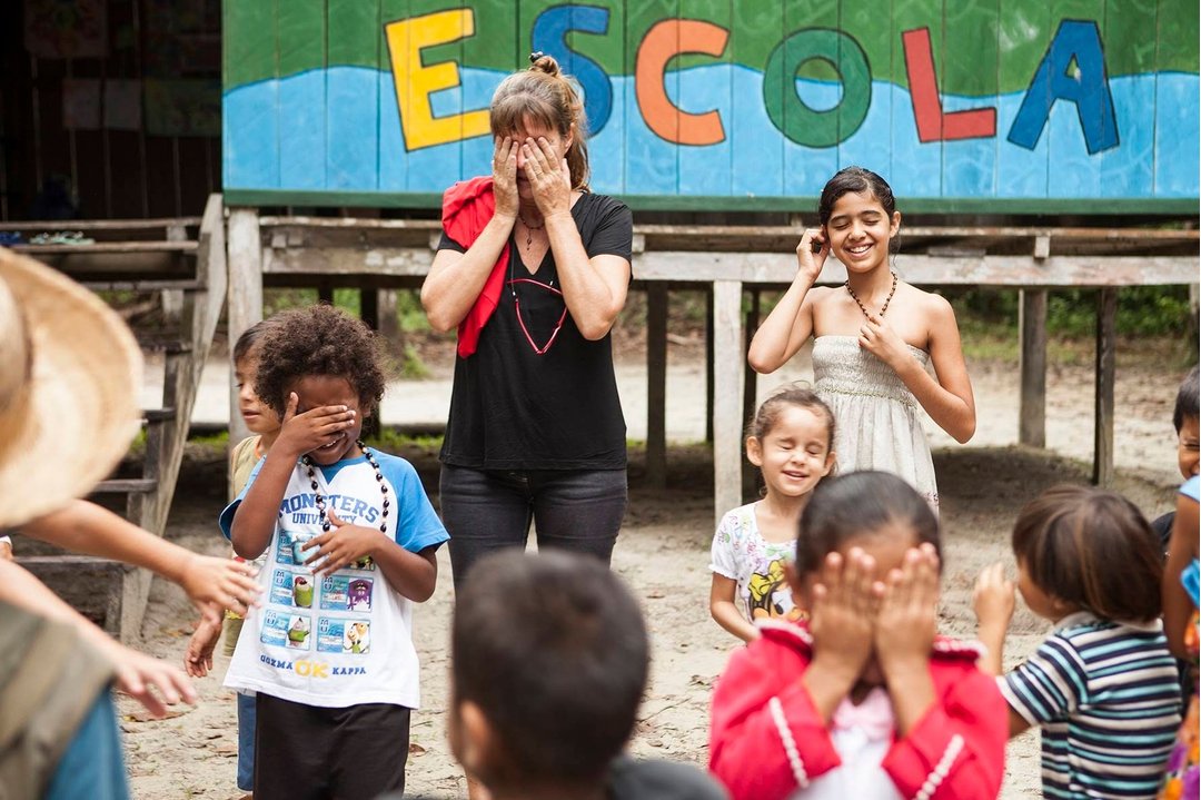 Brincadeira de esconde que une mundos diferentes. Uma viajante e crianças locais brincam de cobrir os olhos em frente a uma escola com a palavra 'ESCOLA' pintada em letras coloridas, durante uma visita comunitária em um roteiro de lodge de luxo na Amazônia brasileira