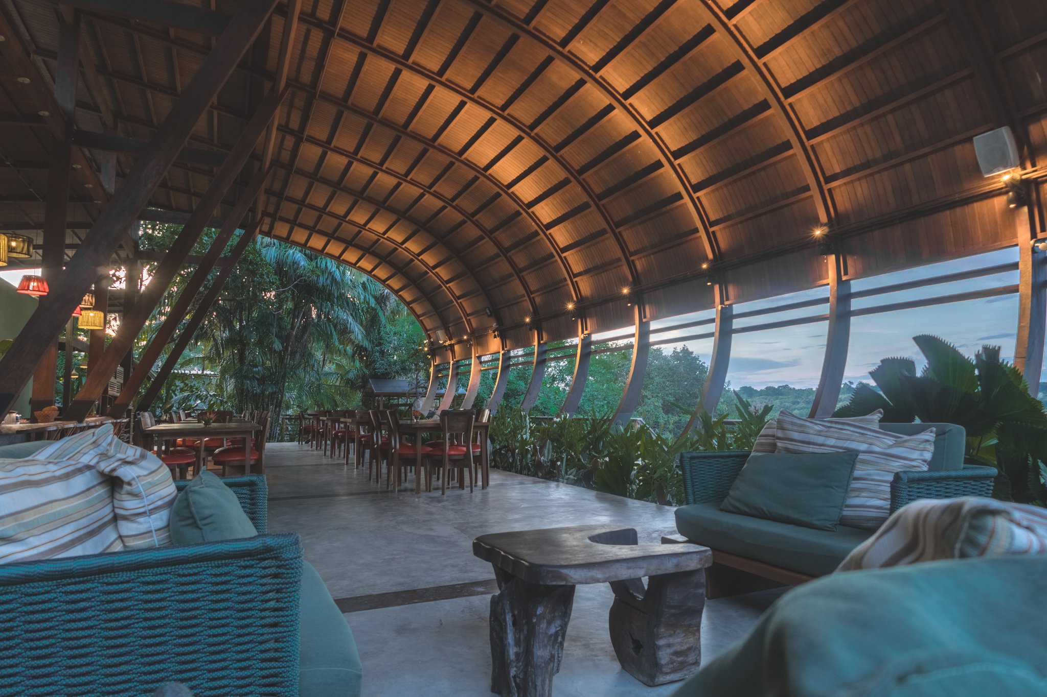 Madeira certificada, floresta como horizonte. Teto abobadado em madeira cobrindo o salão-restaurante aberto de um lodge de luxo na Amazônia brasileira, com sofás de vime, mesa de tronco rústico e vista panorâmica para a copa da floresta ao entardecer