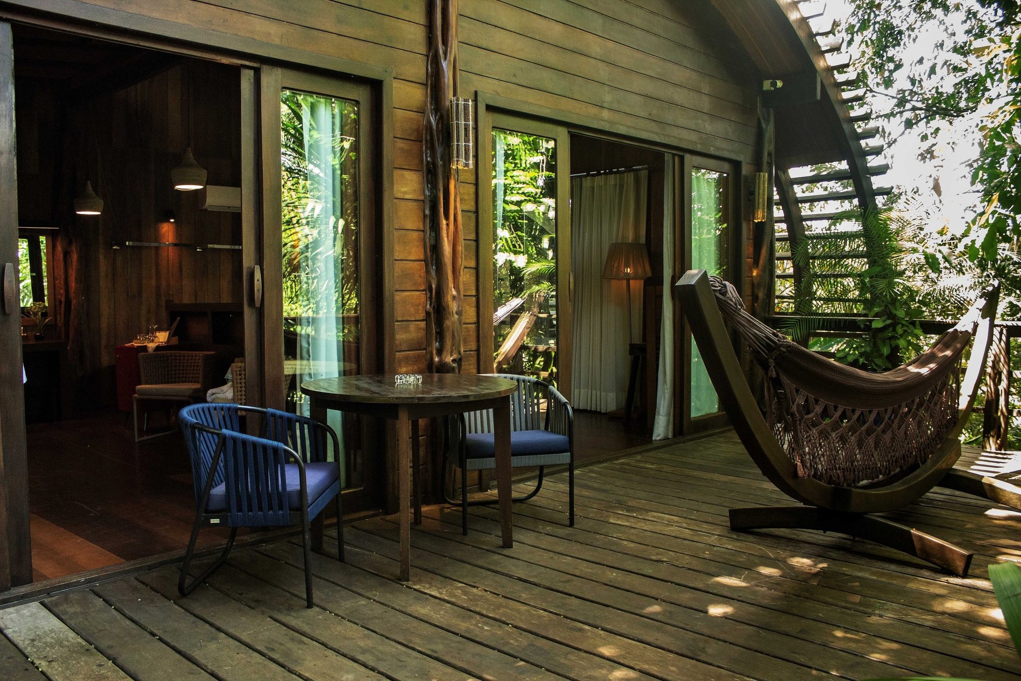 Rede esticada, deck de madeira, mata fechada do lado de fora. Deck de madeira privativo de um lodge de luxo na Amazônia brasileira com mesa redonda, duas cadeiras, rede de descanso em suporte e portas de vidro deslizantes abrindo para uma cabana cercada de floresta