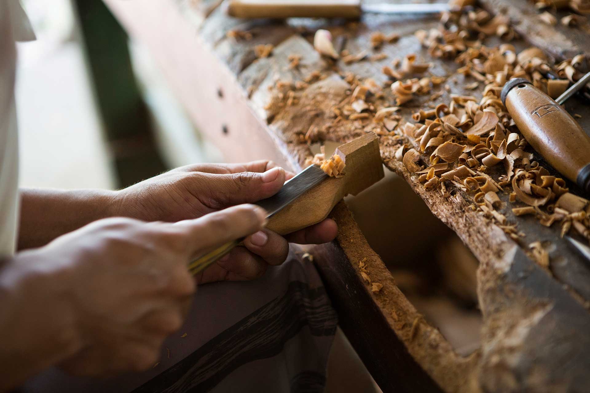 Aparas douradas, mãos firmes — um saber que atravessa gerações. Close das mãos de um artesão utilizando um formão para talhar uma peça de madeira em uma bancada coberta de aparas frescas, representando o artesanato local próximo a um lodge de luxo na Amazônia brasileira