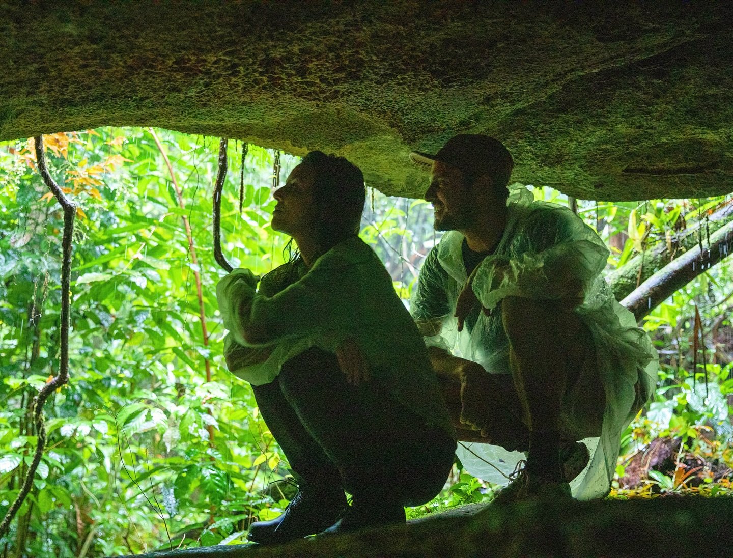 Pedra acima, chuva ao redor — a floresta guarda o que a vista não alcança. Dois viajantes agachados sob uma rocha coberta de musgo, envoltos pela vegetação densa da floresta amazônica em trilha para grutas