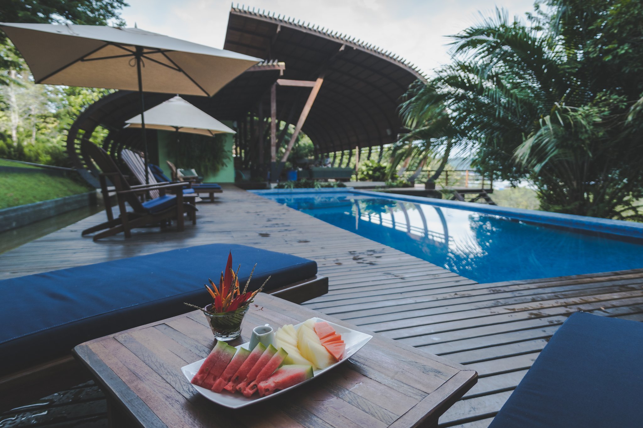 Frutas frescas à beira da piscina, sabores da Amazônia no seu melhor. Uma bandeja de frutas frescas — melancias e melão fatiados — sobre a mesa de uma espreguiçadeira à beira da piscina de um lodge de luxo na Amazônia brasileira, com um pavilhão de madeira curvilíneo e sombrinhas ao fundo