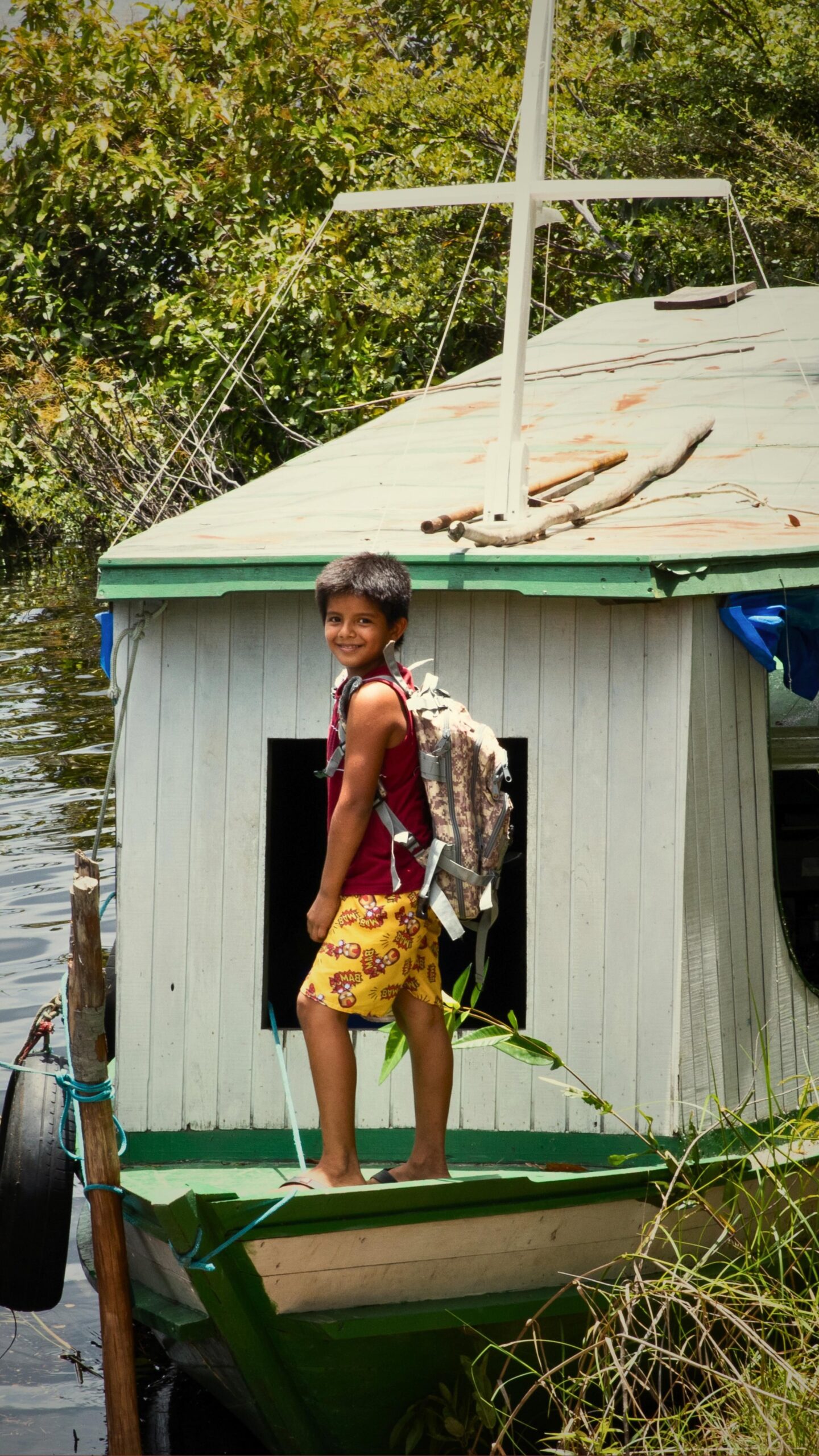 Mochila nas costas, sorriso largo — a Amazônia começa cedo. Um menino sorridente com uma mochila nas costas está sobre o convés de um pequeno barco de madeira atracado entre vegetação exuberante, retratando o acolhimento das comunidades ribeirinhas próximas a um luxury amazon lodge no Brasil