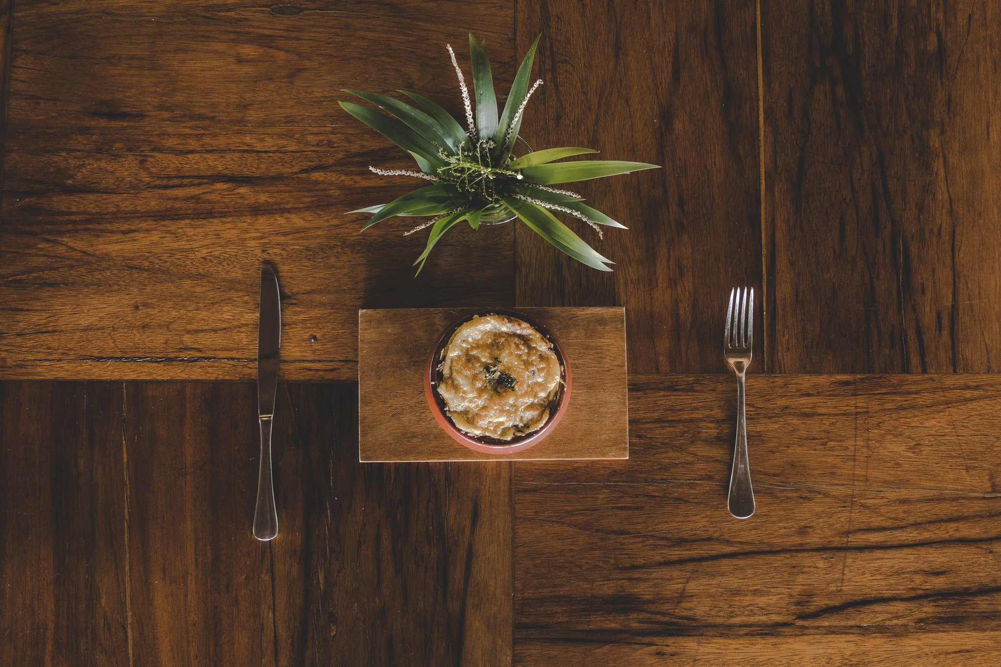 Ingredientes simples, apresentados com elegância na Amazônia. Vista aérea de um prato gratinado em ramequin de cerâmica sobre uma tábua de madeira com faca e garfo, parte da experiência gastronômica de um lodge de luxo na Amazônia brasileira