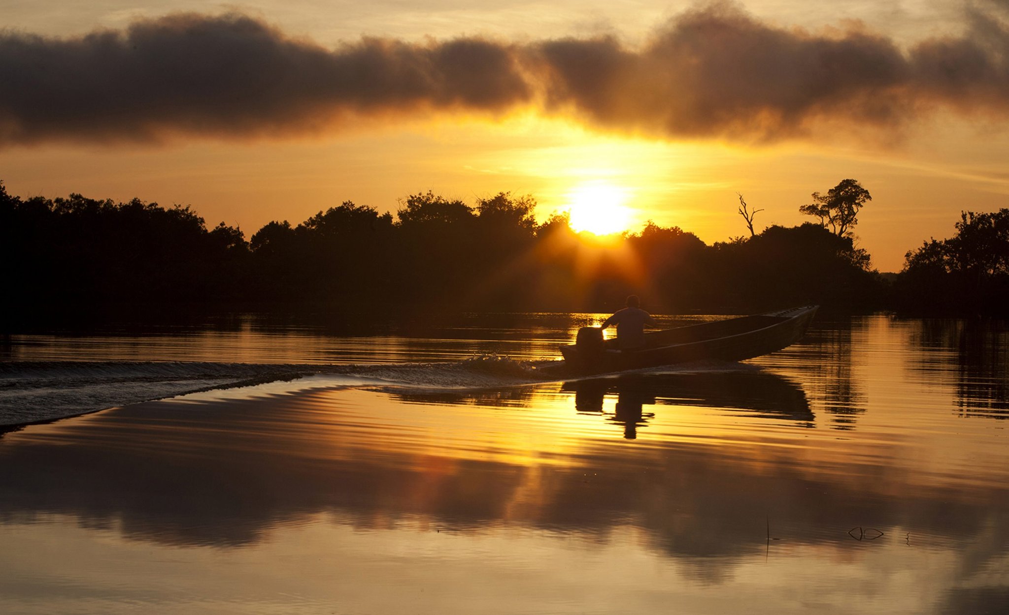 Voadeira, águas negras e um céu que pega fogo. Uma voadeira em silhueta desliza sobre um rio espelhado em direção a um pôr do sol dourado, com a mata escura refletida nas águas calmas do Rio Negro