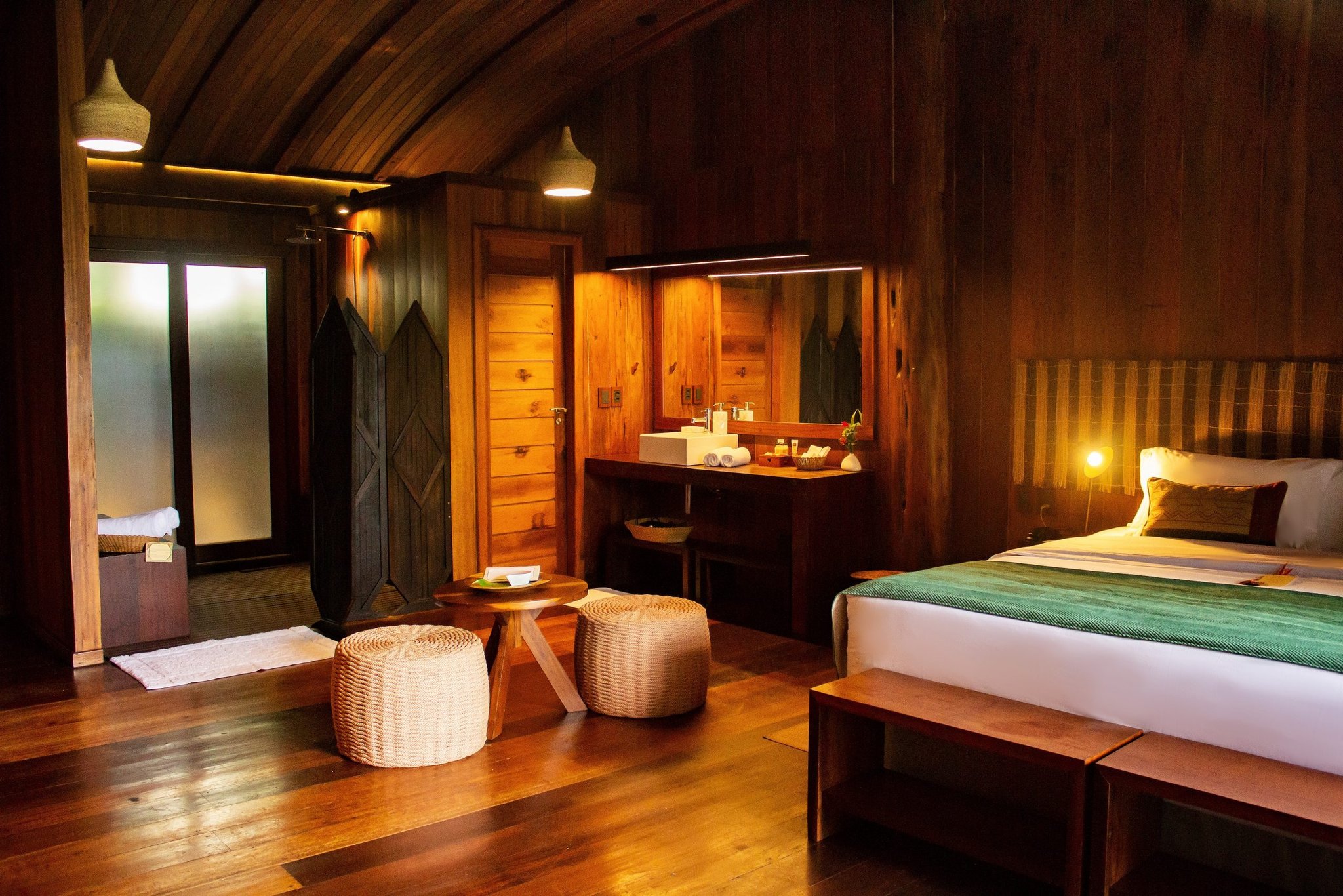 Rich hardwood, soft lamplight, wicker accents — comfort deep in the rainforest. Warmly lit timber cabin interior at a luxury amazon lodge brazil, featuring a queen bed with green throw, wicker ottomans, open vanity with white basin, and polished hardwood floors beneath an arched wooden ceiling