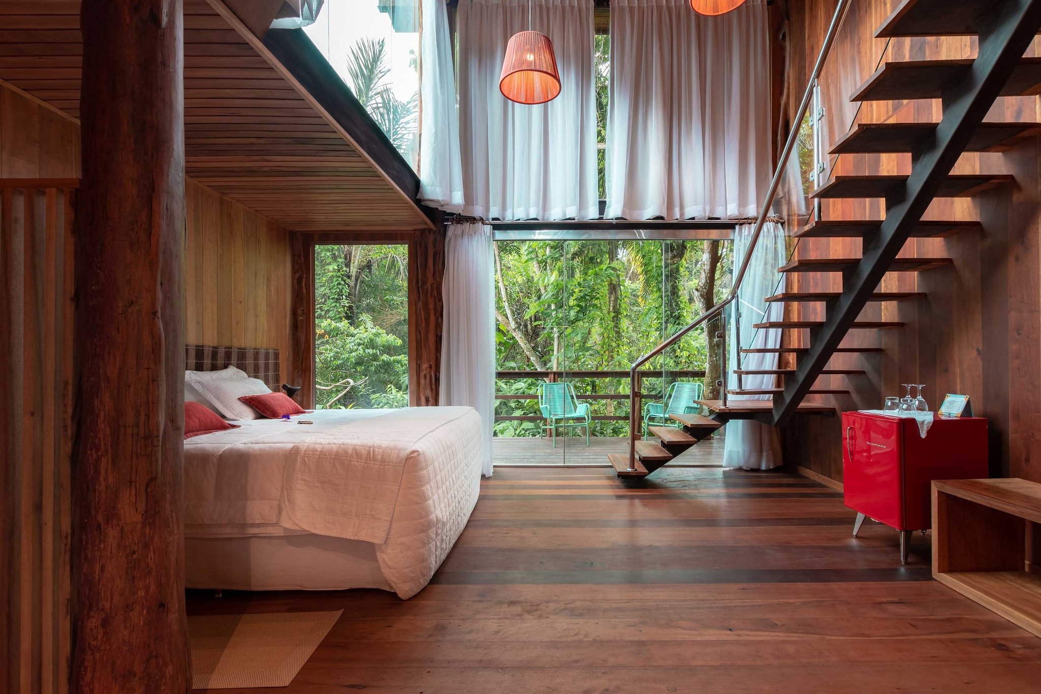 Madeira nobre, cortinas leves e floresta amazônica a dois passos — o chalé-árvore que convida a ficar. Quarto em mezanino de um lodge de luxo na Amazônia brasileira, com paredes e piso de madeira certificada, cama queen com almofadas vermelhas, escada flutuante em aço, frigobar retrô vermelho e portas de vidro do piso ao teto abrindo para um deck rodeado de mata fechada