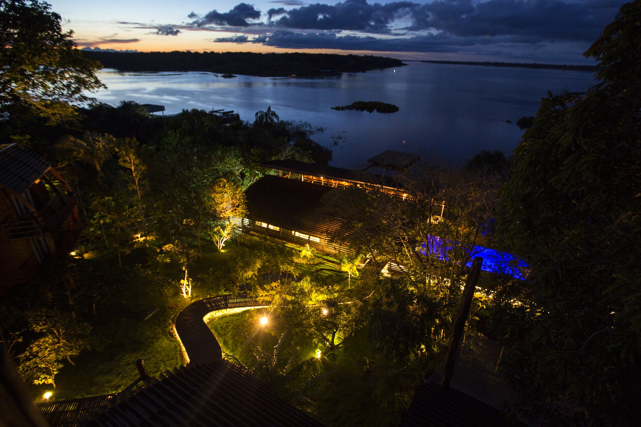 Luzes quentes no coração da floresta, o rio negro se estende até o horizonte. Vista aérea ao entardecer de um lodge de luxo na Amazônia brasileira, com edificações de madeira iluminadas por luzes douradas, passarelas sinuosas e jardim exuberante descendo até um amplo rio escuro salpicado de ilhas florestadas sob um céu dramático
