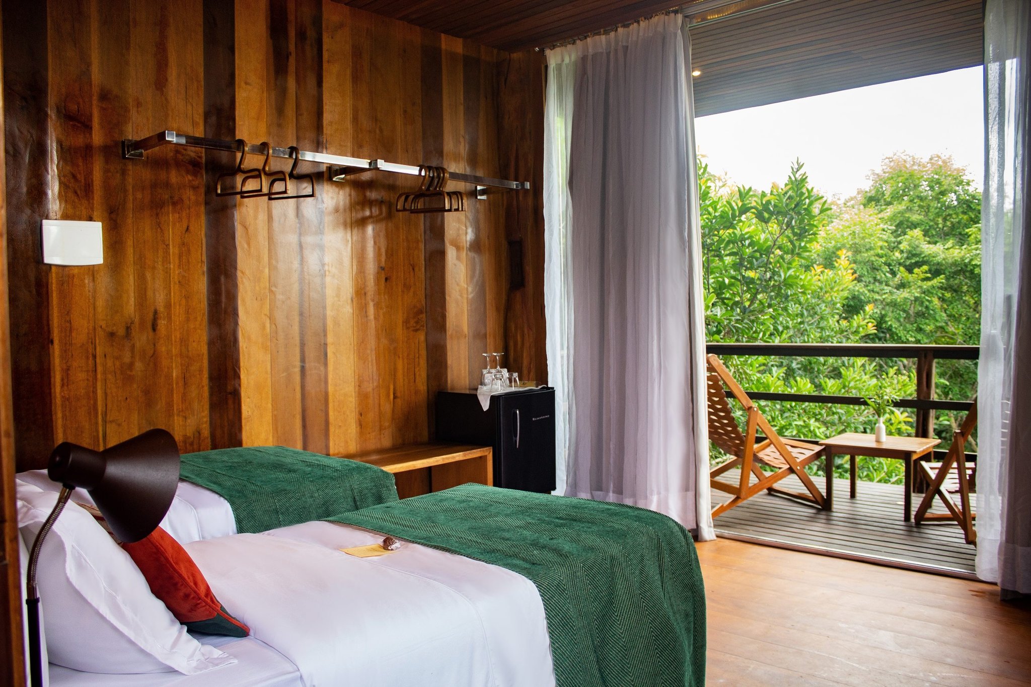 Sleep among the canopy — sheer curtains, warm timber, and a forest balcony all to yourself. Twin beds with green throws in a wood-panelled room at a luxury amazon lodge brazil, opening onto a private balcony with reclining chairs set among lush green treetops