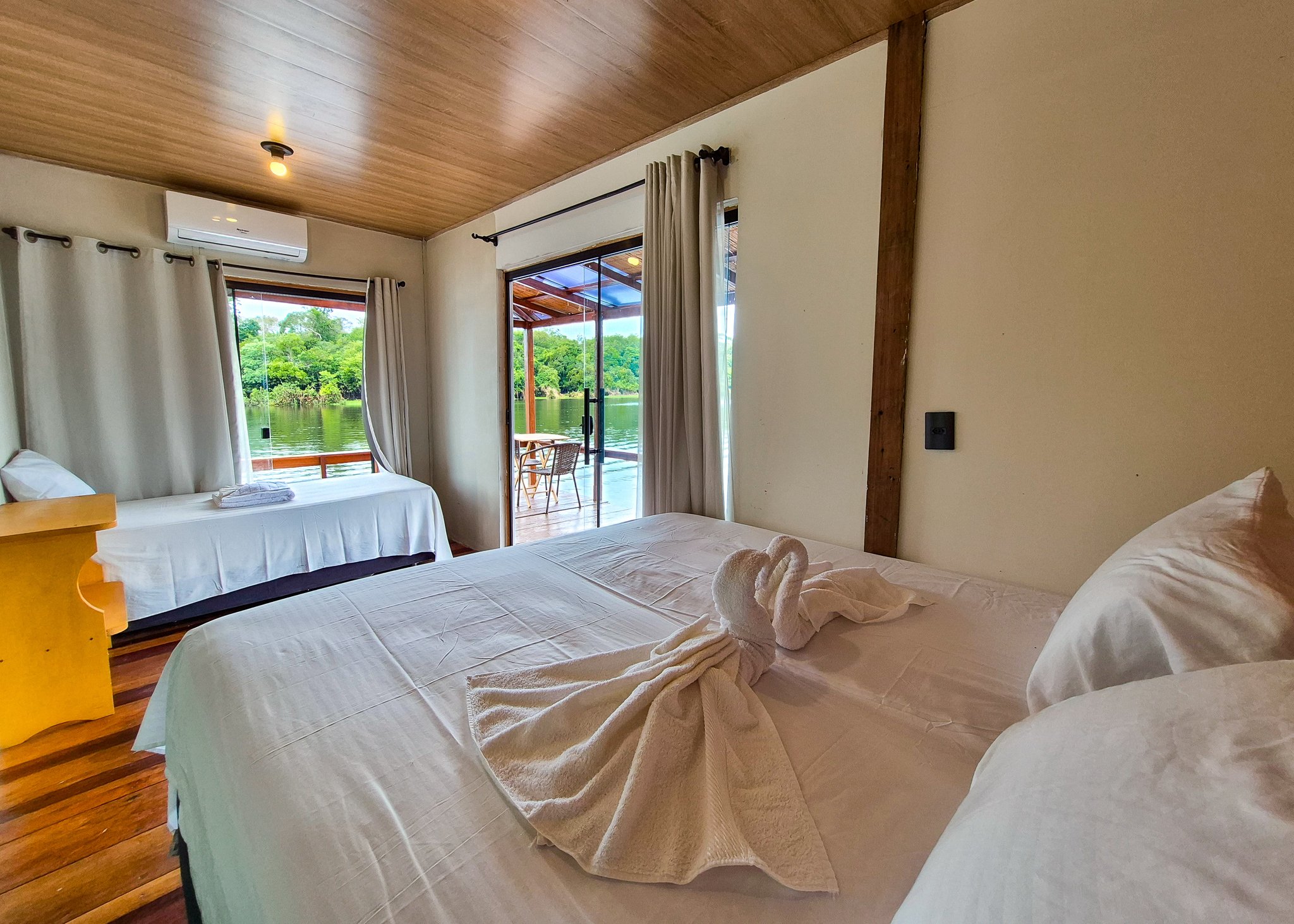 Towel swans, crisp beds, and a rainforest river framed in your doorway. Twin bedroom with towel art on white linen beds, wooden ceiling, air conditioning, and a glass sliding door opening onto a deck with a river and rainforest view — part of a manaus jungle tour eco floating lodge