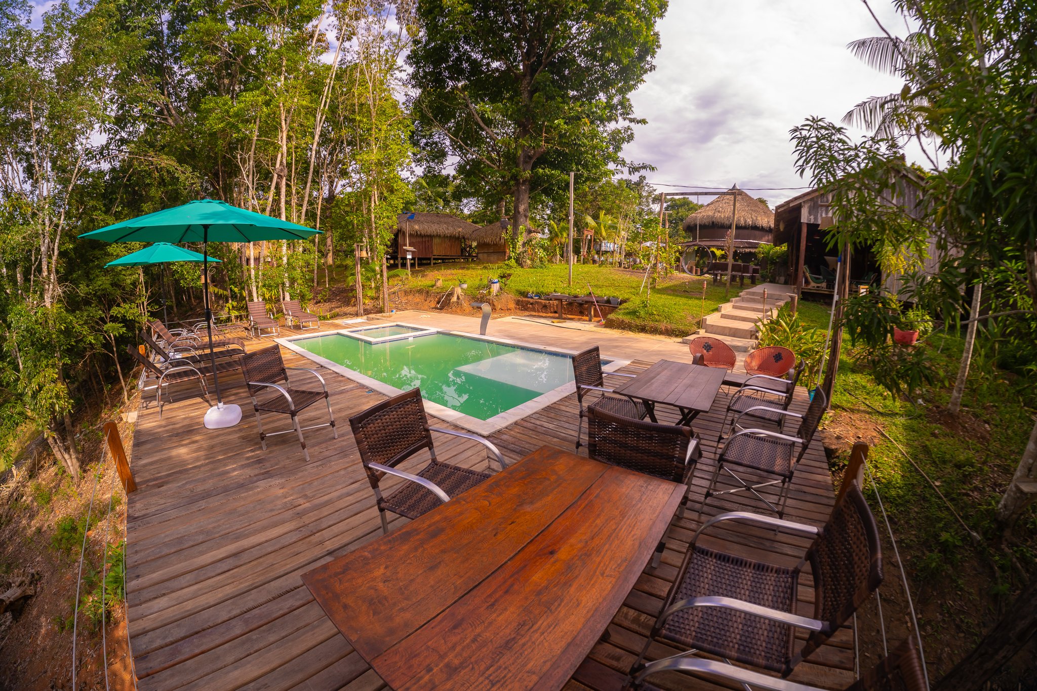 Sundowners on the deck, towering canopy all around. Hardwood deck with dining tables, wicker chairs, a teal umbrella, and a rectangular swimming pool surrounded by rainforest trees and thatched-roof bungalows at a jungle lodge on a manaus jungle tour