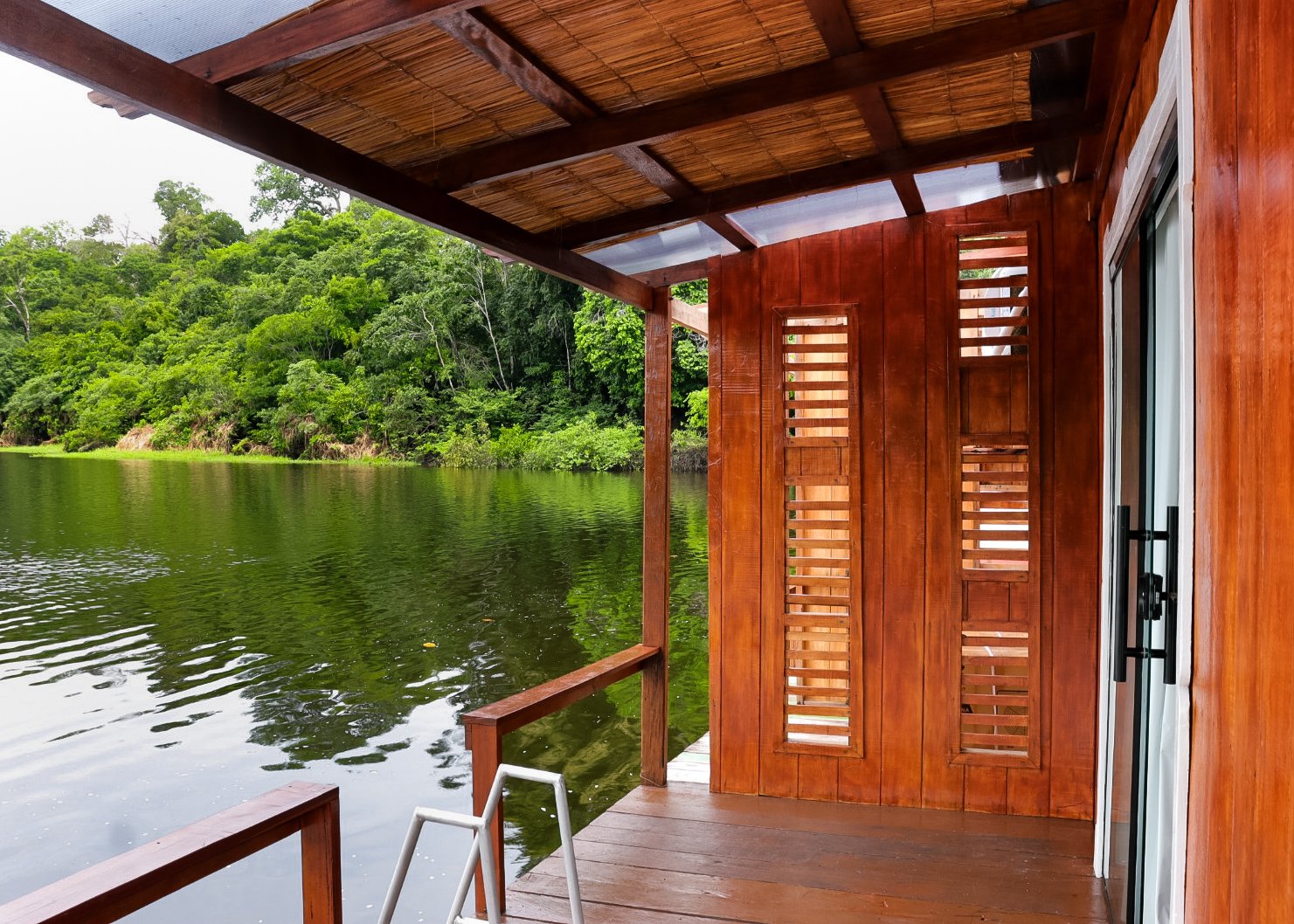 Still water, deep green canopy — your private deck at the edge of the Amazon. Covered timber deck of a jungle lodge on a manaus jungle tour, with louvred hardwood shutters and a thatched ceiling opening onto a calm dark-water channel bordered by dense rainforest