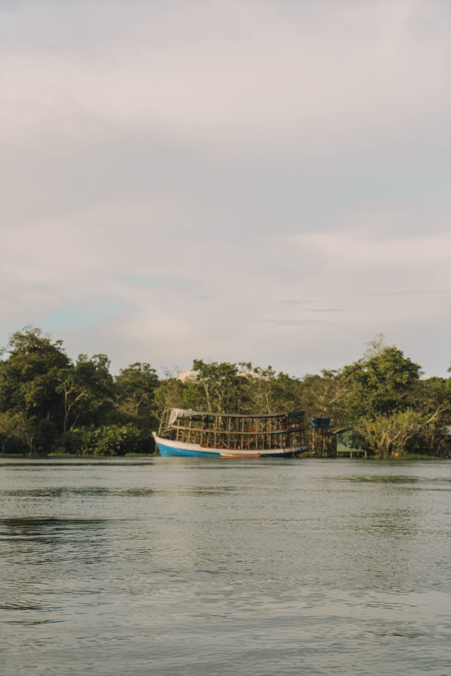 A blue-and-white wooden river boat with an open timber frame upper deck moored along a lush tropical riverbank on a manaus jungle tour