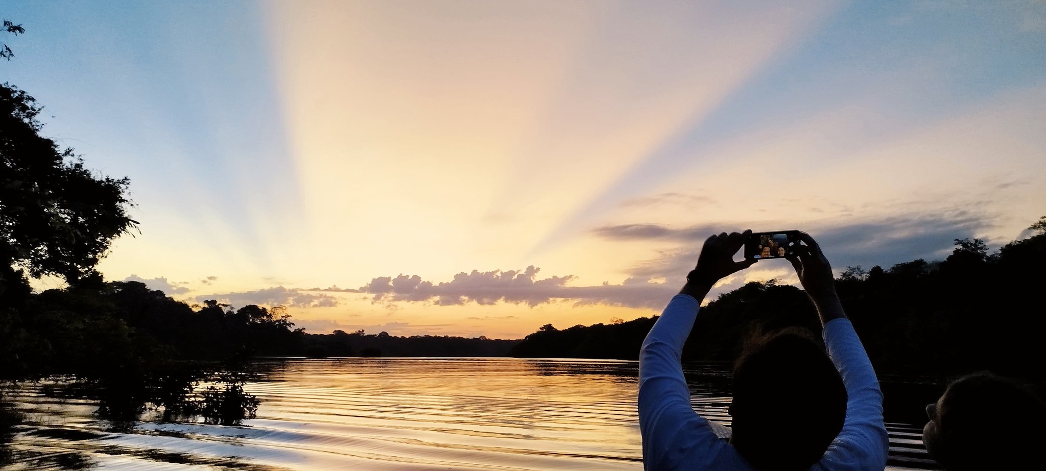 Crepuscular rays, golden water, and one phone that cannot put itself down. A traveller holds up a smartphone to photograph a dramatic sunset over a calm jungle river, with crepuscular rays fanning out across the sky on a manaus jungle tour