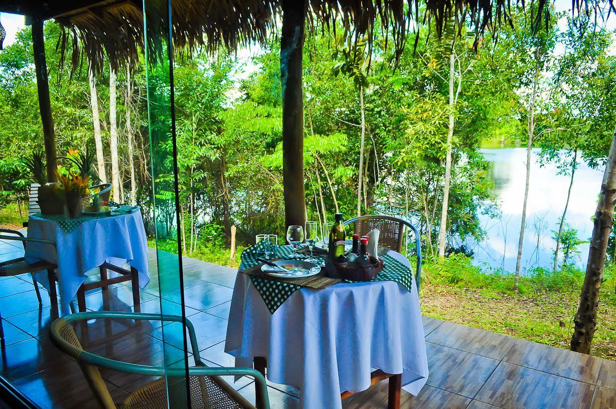 Dinner with a view — still water, tall trees, and a bottle of wine waiting. Open-air thatched dining terrace on a manaus jungle tour, with white-clothed tables set for two, wine bottles, and a tranquil forest lake visible through the trees