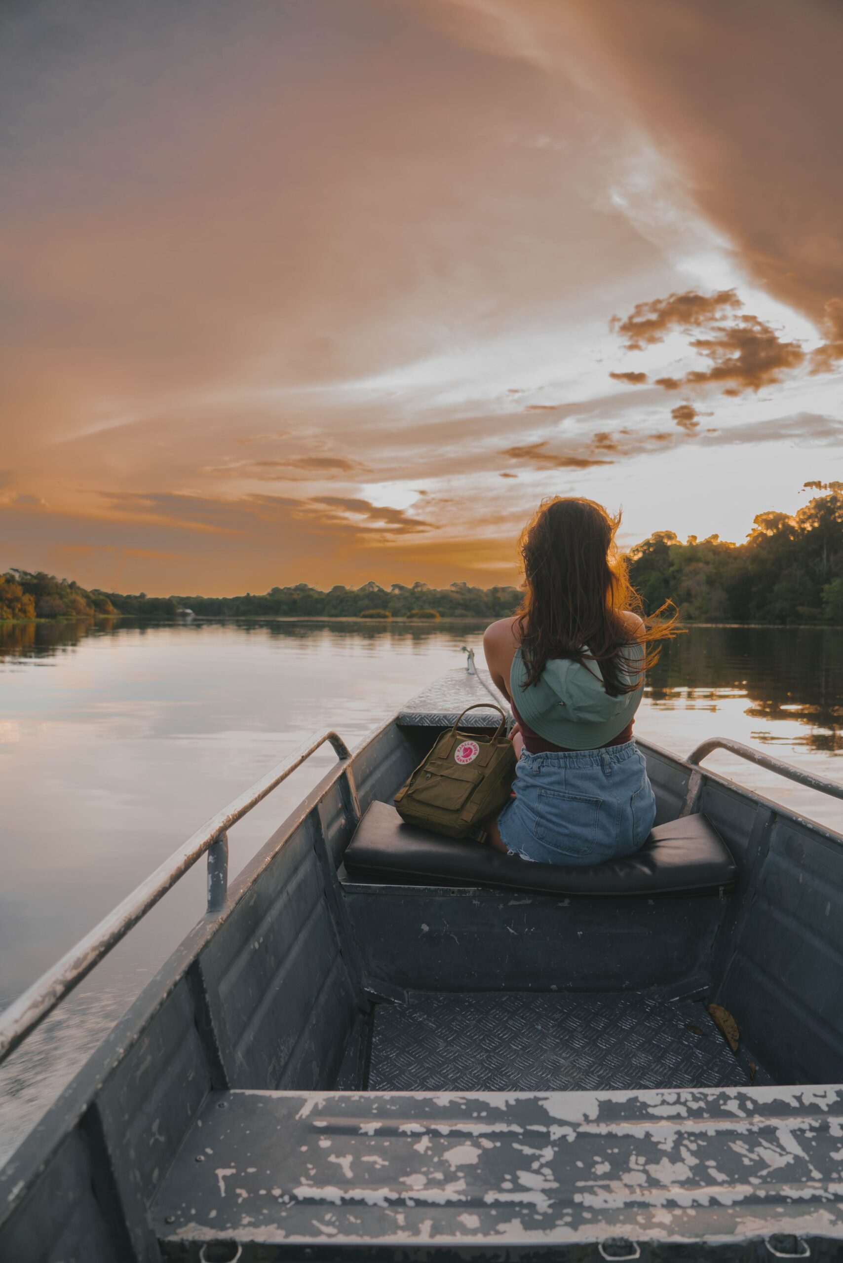 Golden hour on the river — hair catching the breeze, nothing ahead but open water and burning sky. Photo: Yuri Panichi A traveller sits at the bow of a small aluminium boat on a glassy jungle river, watching a vivid orange sunset during a manaus jungle tour, dense rainforest lining both banks